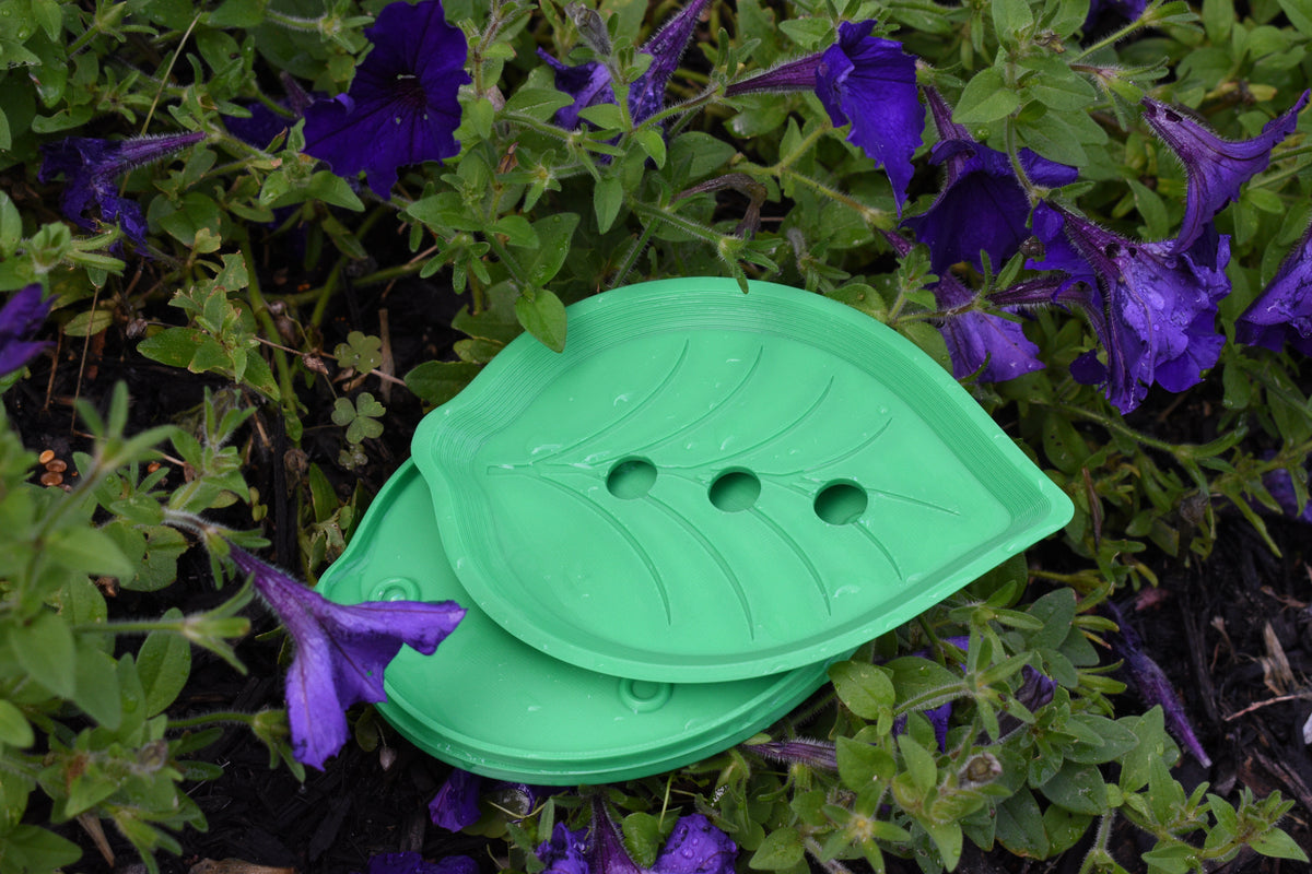 Top-down view of the green 3D-printed FloraDrain self-draining soap dish from the Solara collection with its catch tray, resting outdoors among green leaves and purple flowers.