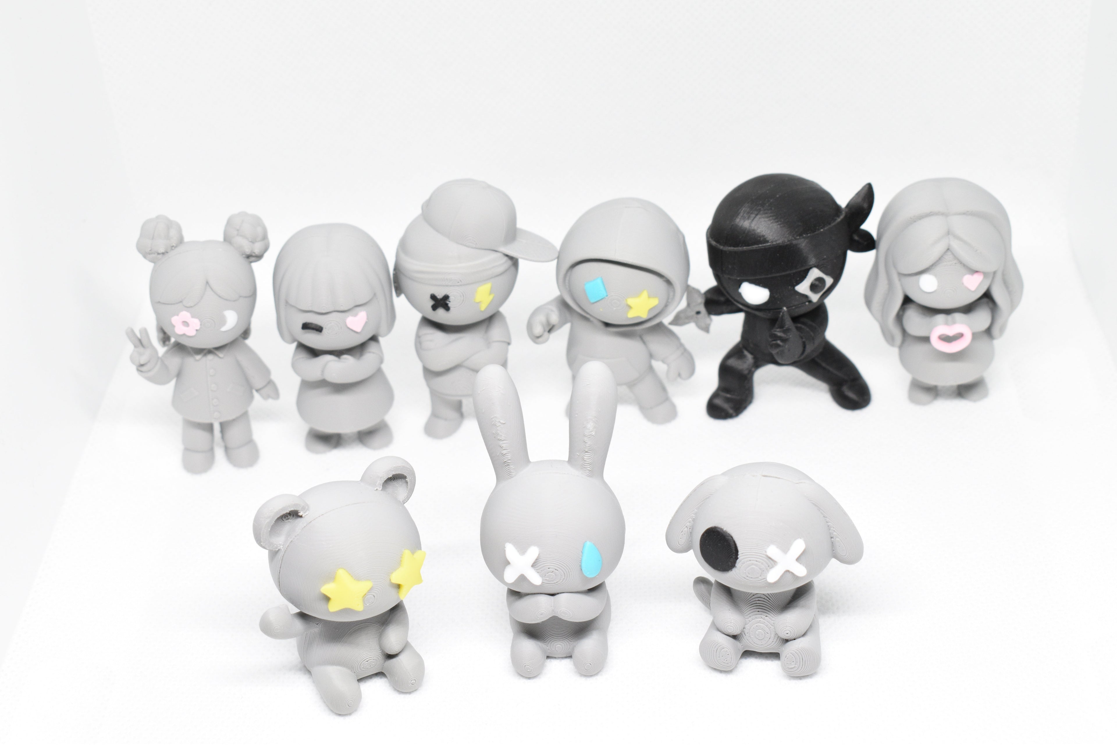 Nine gray Monomoodz figurines with colorful facial accents arranged in two rows on a white surface.
