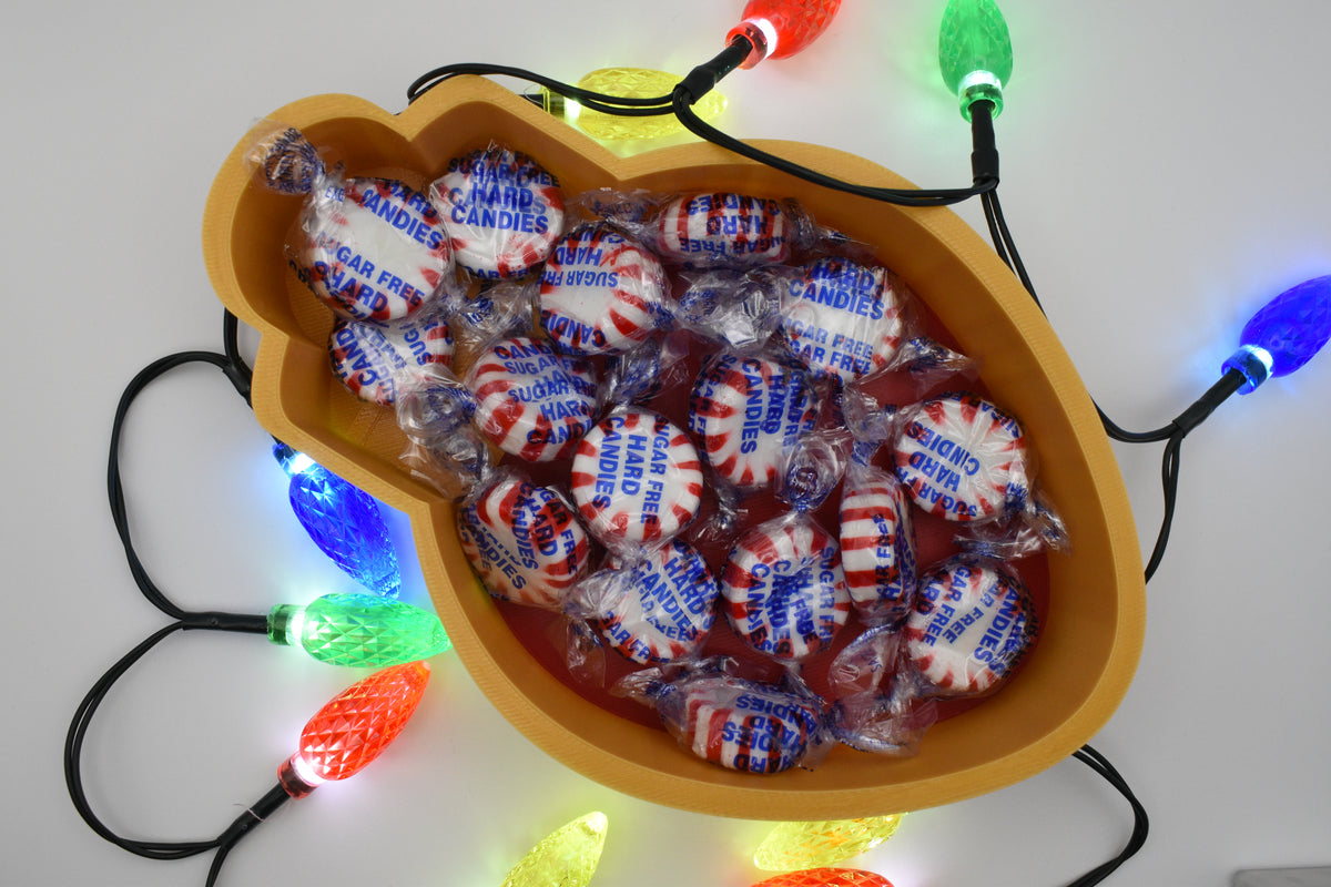 Overhead view of the Layertreat BulbBowl 3D printed candy dish filled with wrapped peppermint candies and framed by colorful glowing Christmas lights.