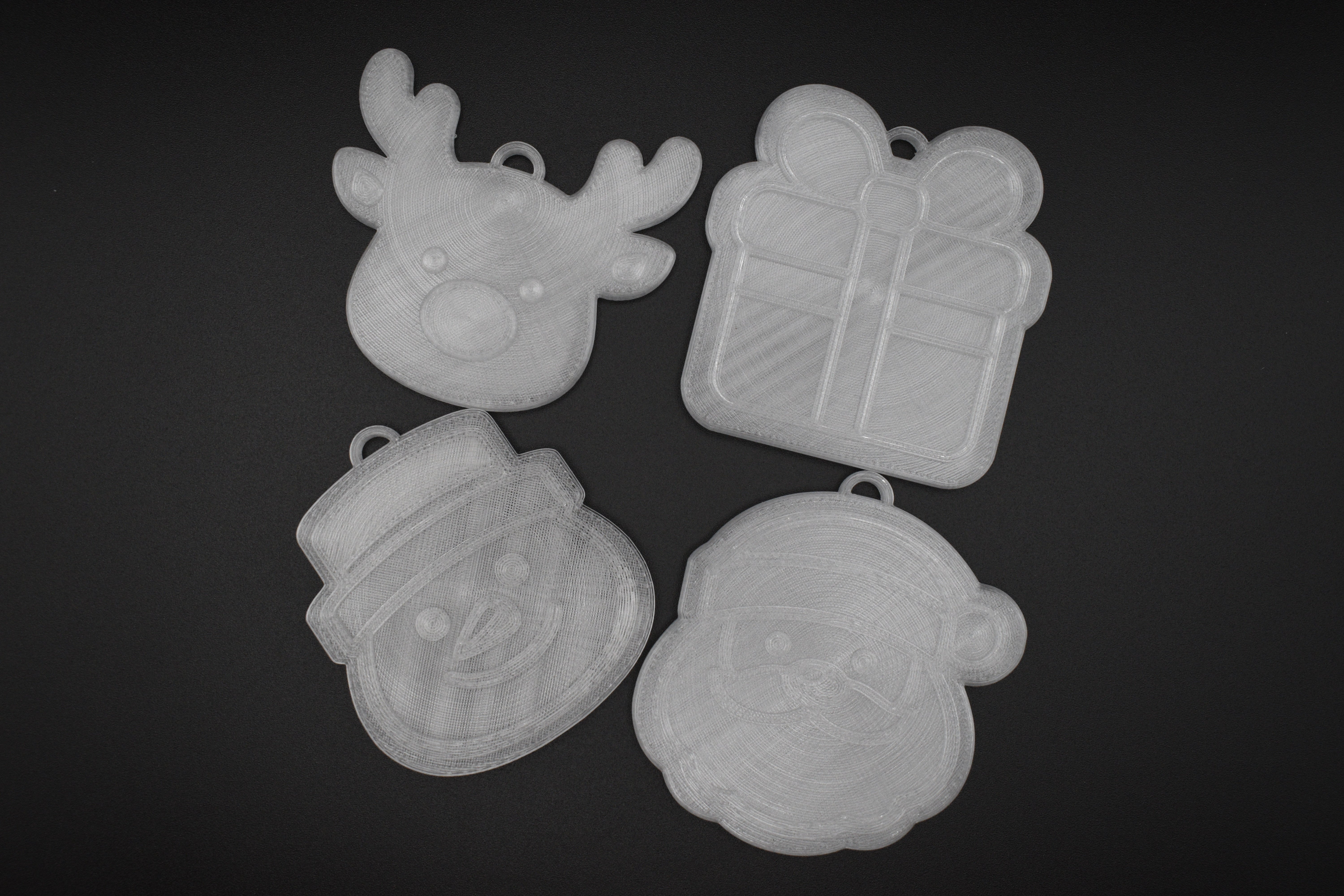 Flat lay group photo of the 4-piece 3D printed translucent Christmas ornament set including reindeer, present, snowman, and Santa Claus on a dark surface.