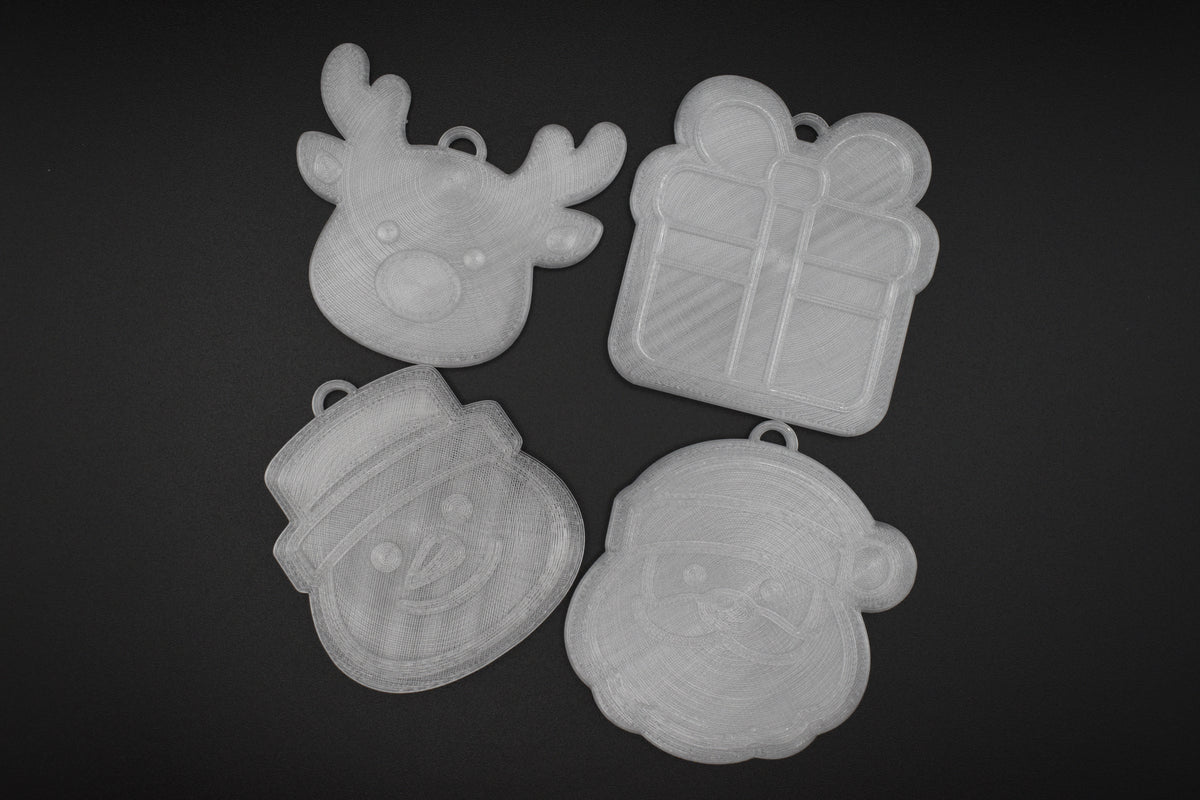 Flat lay group photo of the 4-piece 3D printed translucent Christmas ornament set including reindeer, present, snowman, and Santa Claus on a dark surface.