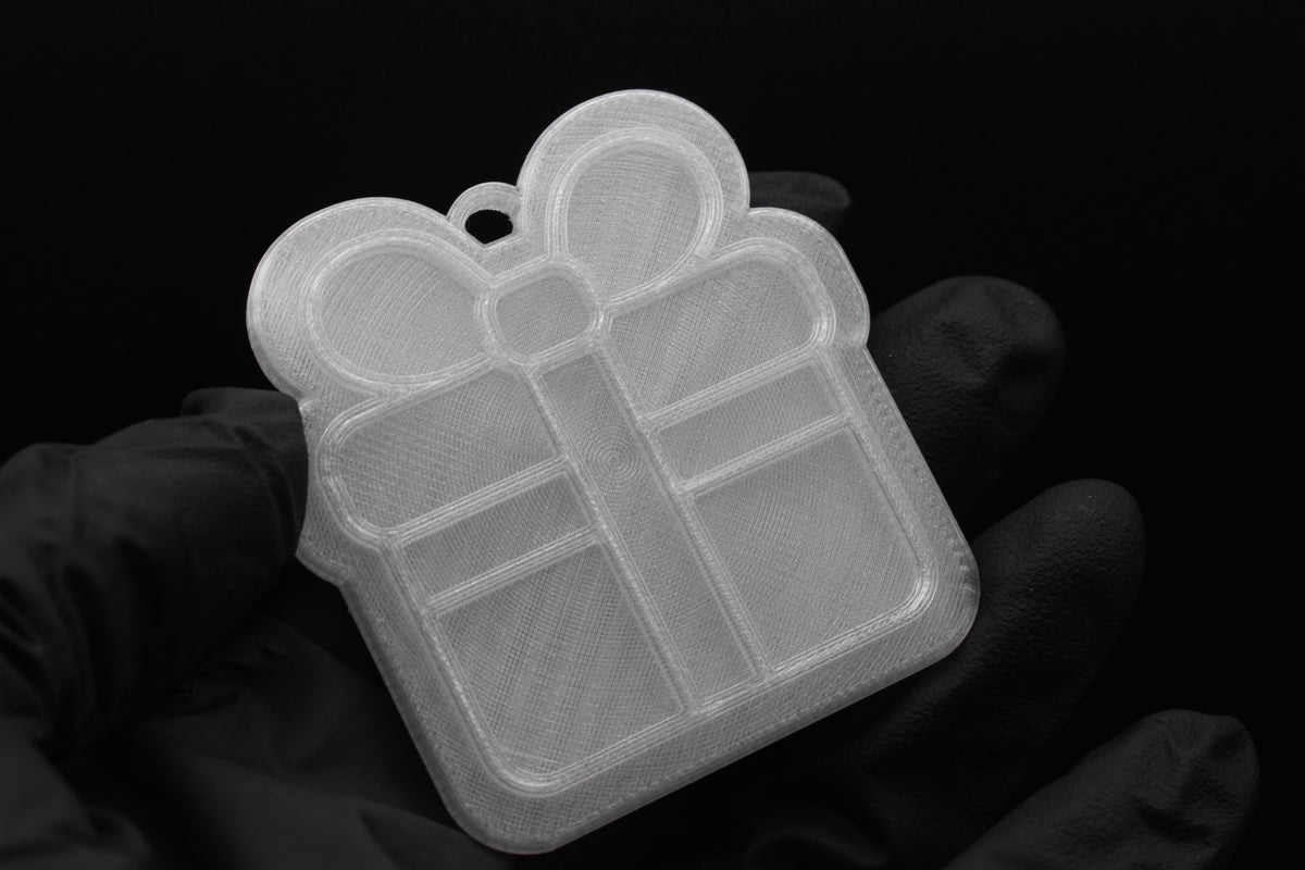 Handheld studio shot of a translucent 3D printed gift box present ornament featuring a ribbon bow detail on a black background.