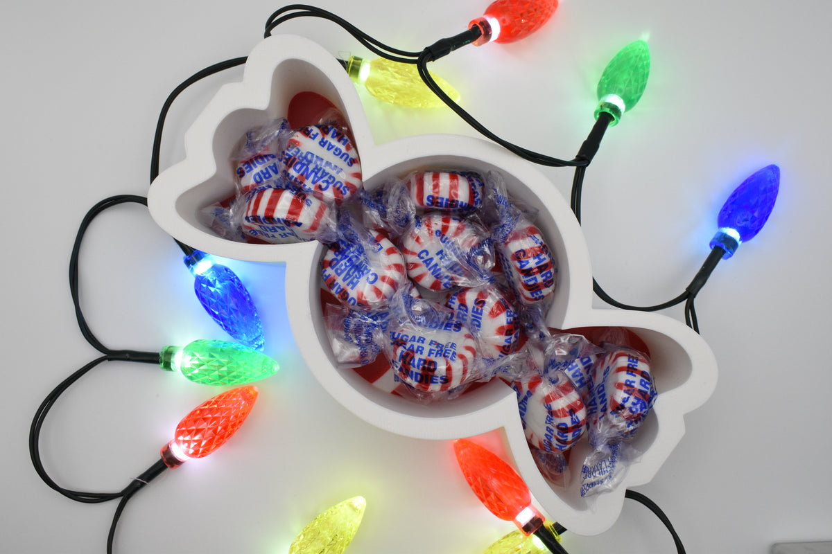 Overhead shot of the Layertreat 3D printed peppermint candy dish filled with wrapped peppermint candies, framed by colorful glowing holiday lights.