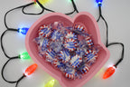 Overhead shot of the pink Layertreat MittenBowl 3D printed candy dish filled with wrapped peppermint candies, framed by colorful glowing holiday lights.