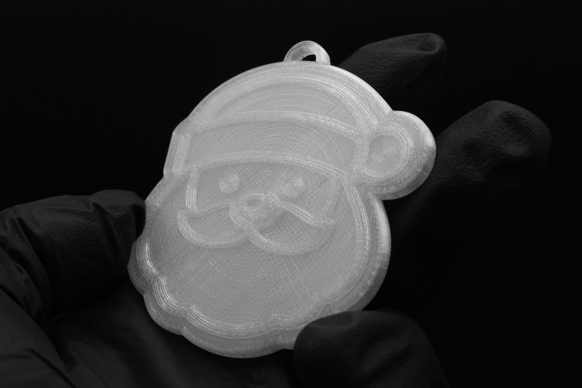 Handheld view of a 3D printed translucent clear Santa Claus Christmas tree ornament showing detailed facial features, hat, and beard against a dark background.
