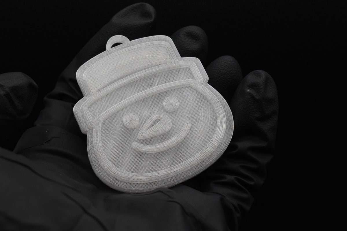 Handheld view of a 3D printed clear translucent snowman Christmas ornament with top hat detail against a dark background.