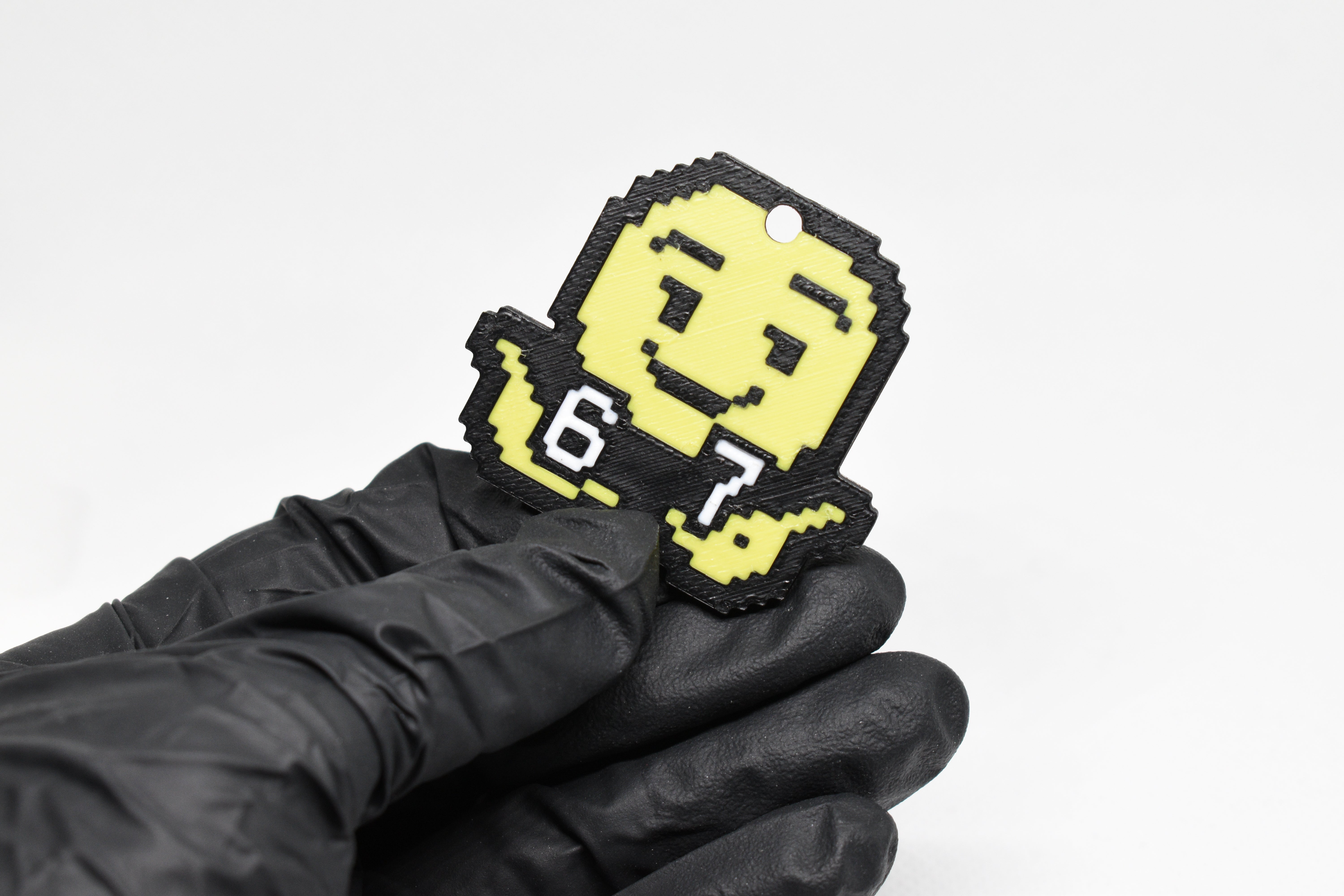 Pixelated 67 emoji keychain held in a gloved hand, showing yellow face and raised arms.