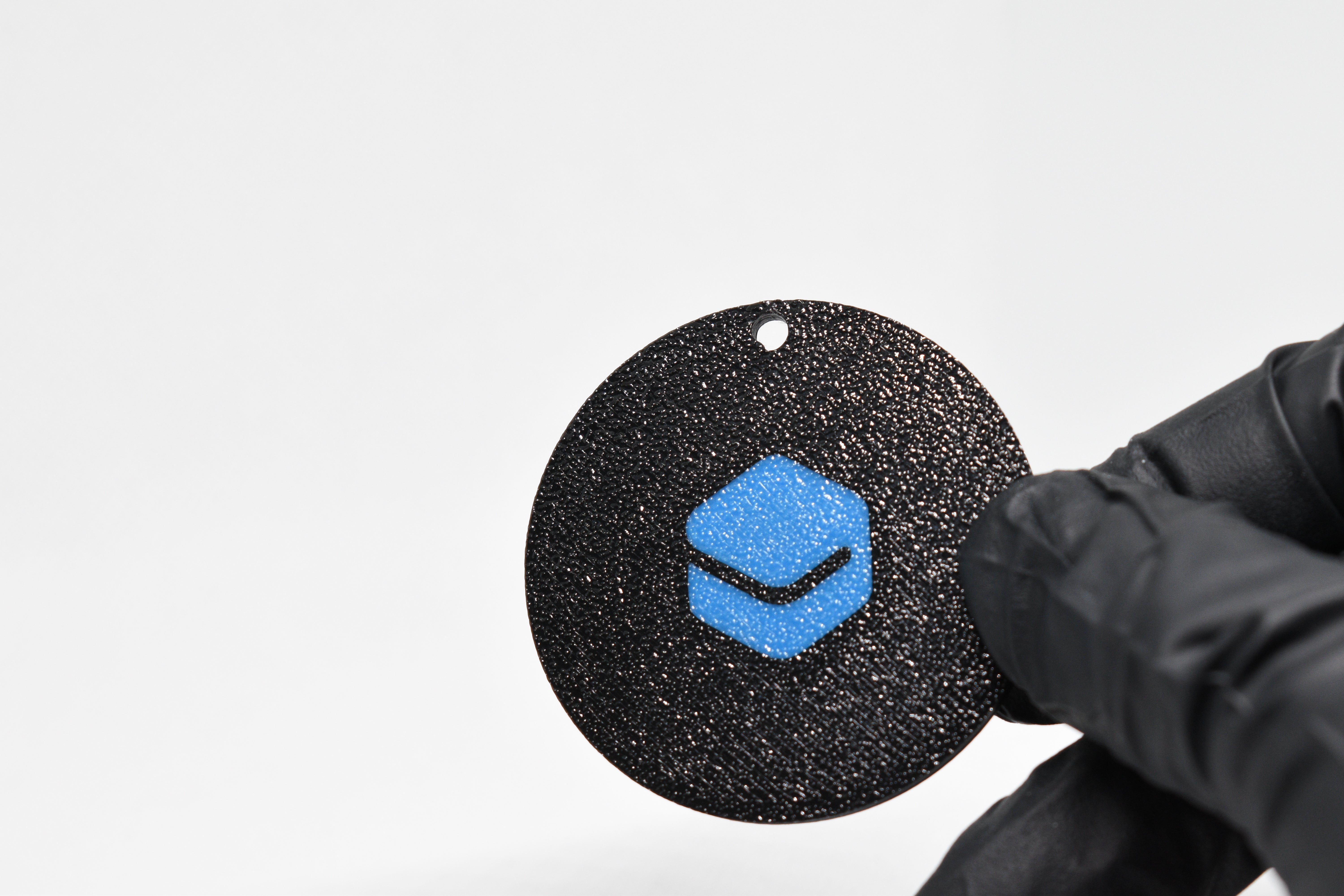 Black textured round object with a blue Layerplay logo held by a gloved hand on a white background