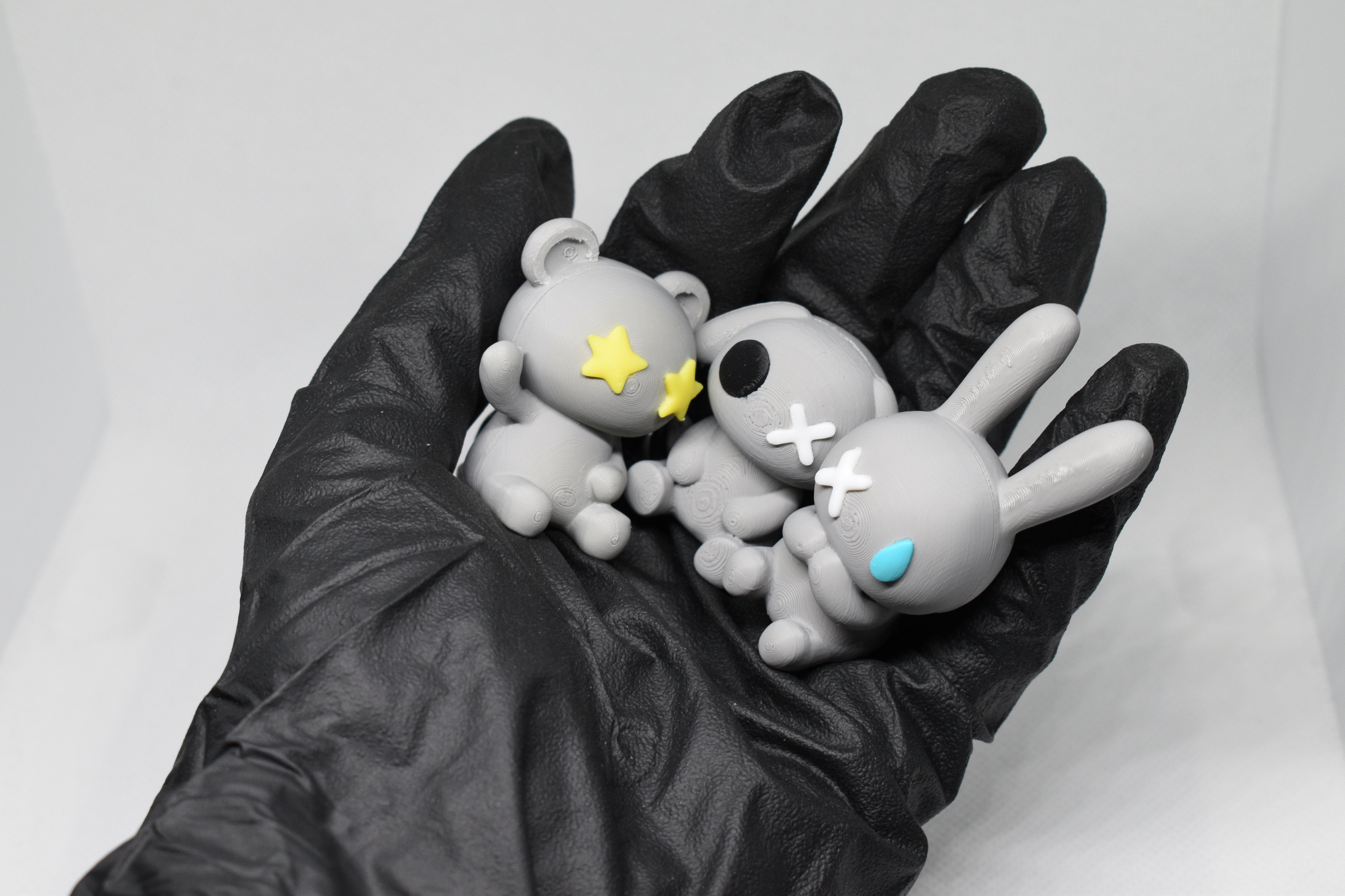 Three Monomoodz figurines held in a gloved hand, featuring bear, skull, and bunny designs on a white background.