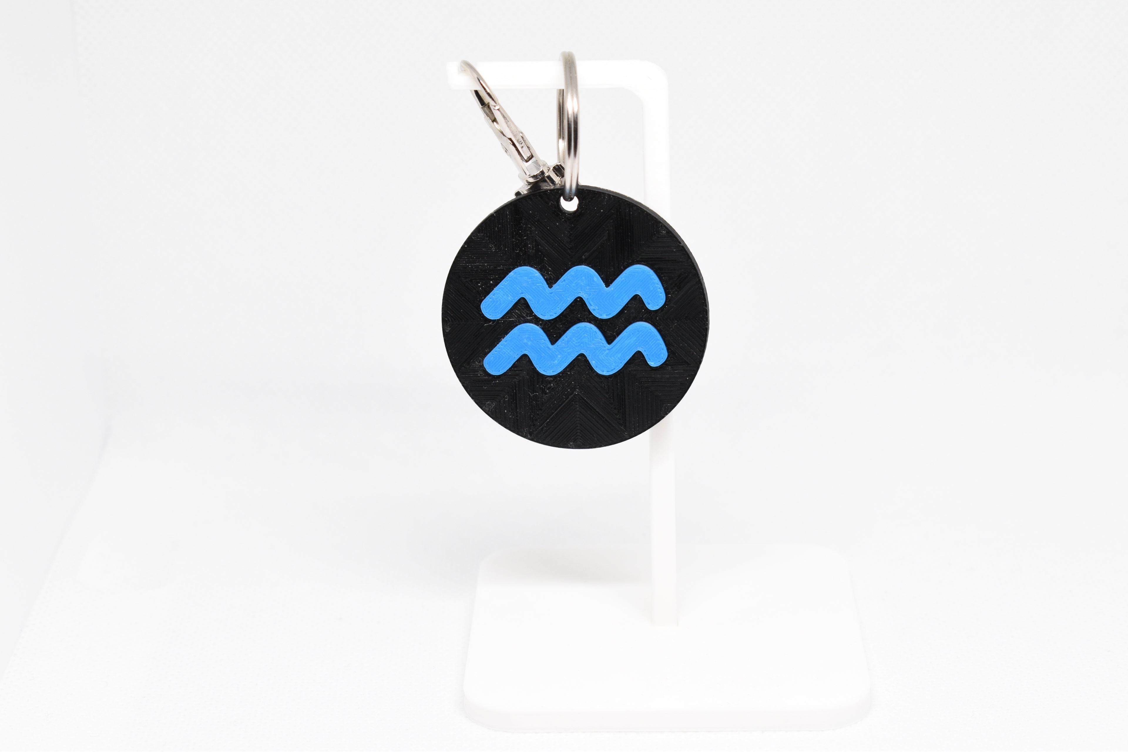 Aquarius zodiac keychain with blue symbol displayed on a white stand with metal ring and clip.