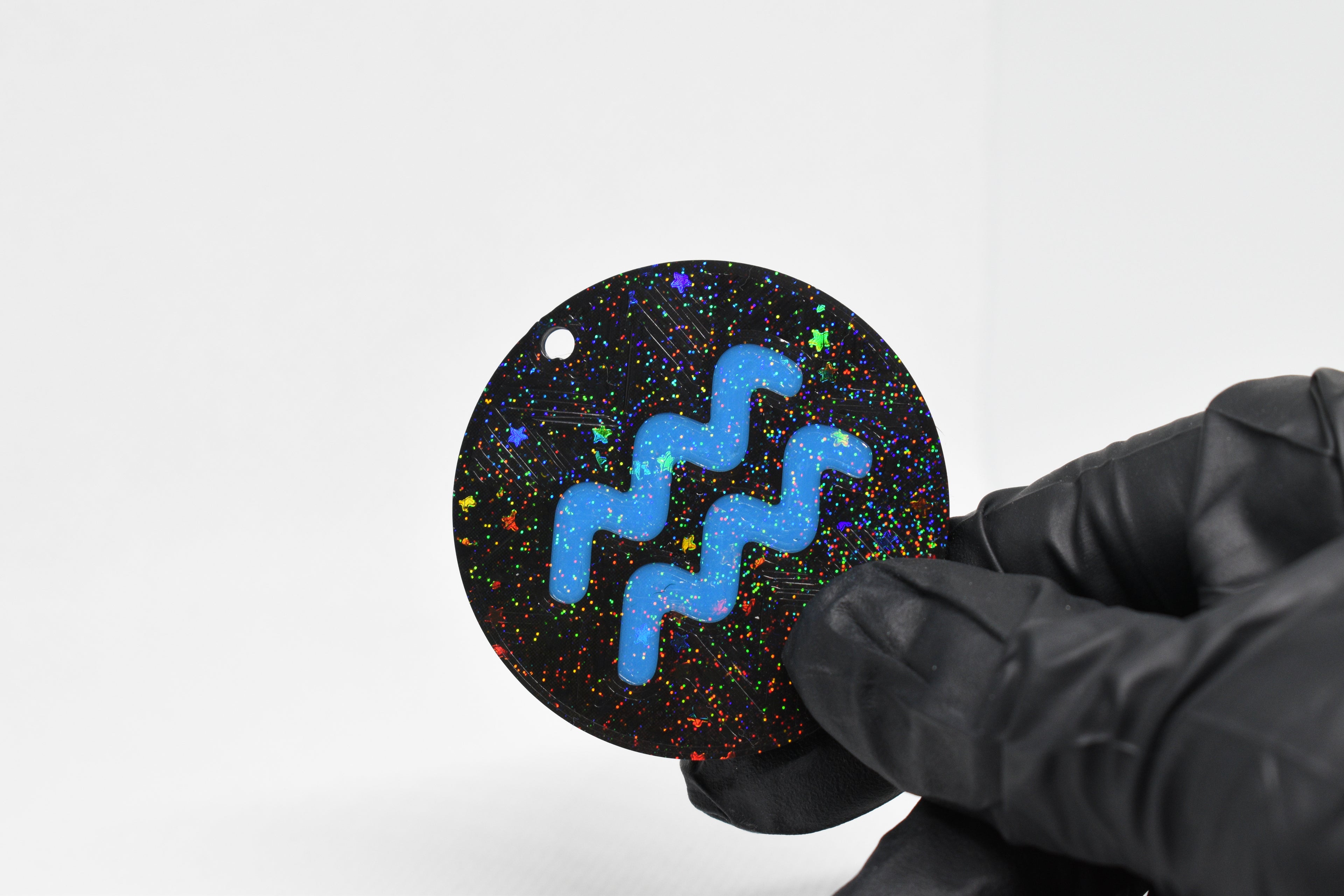 Aquarius zodiac keychain with blue symbol and glitter‑black cosmic background held in a gloved hand.