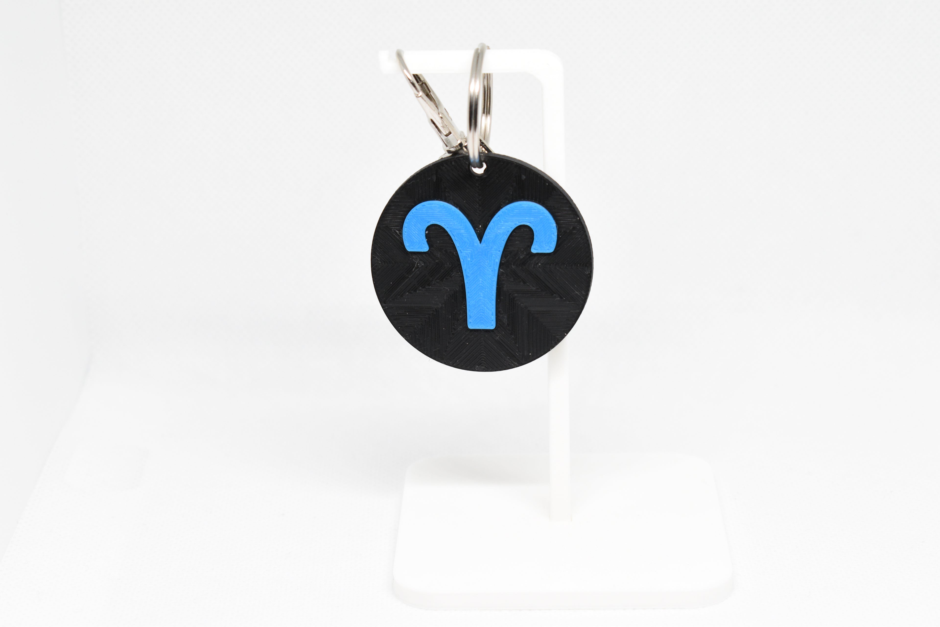 Aries zodiac keychain with blue symbol displayed on a white stand with metal ring and clip.