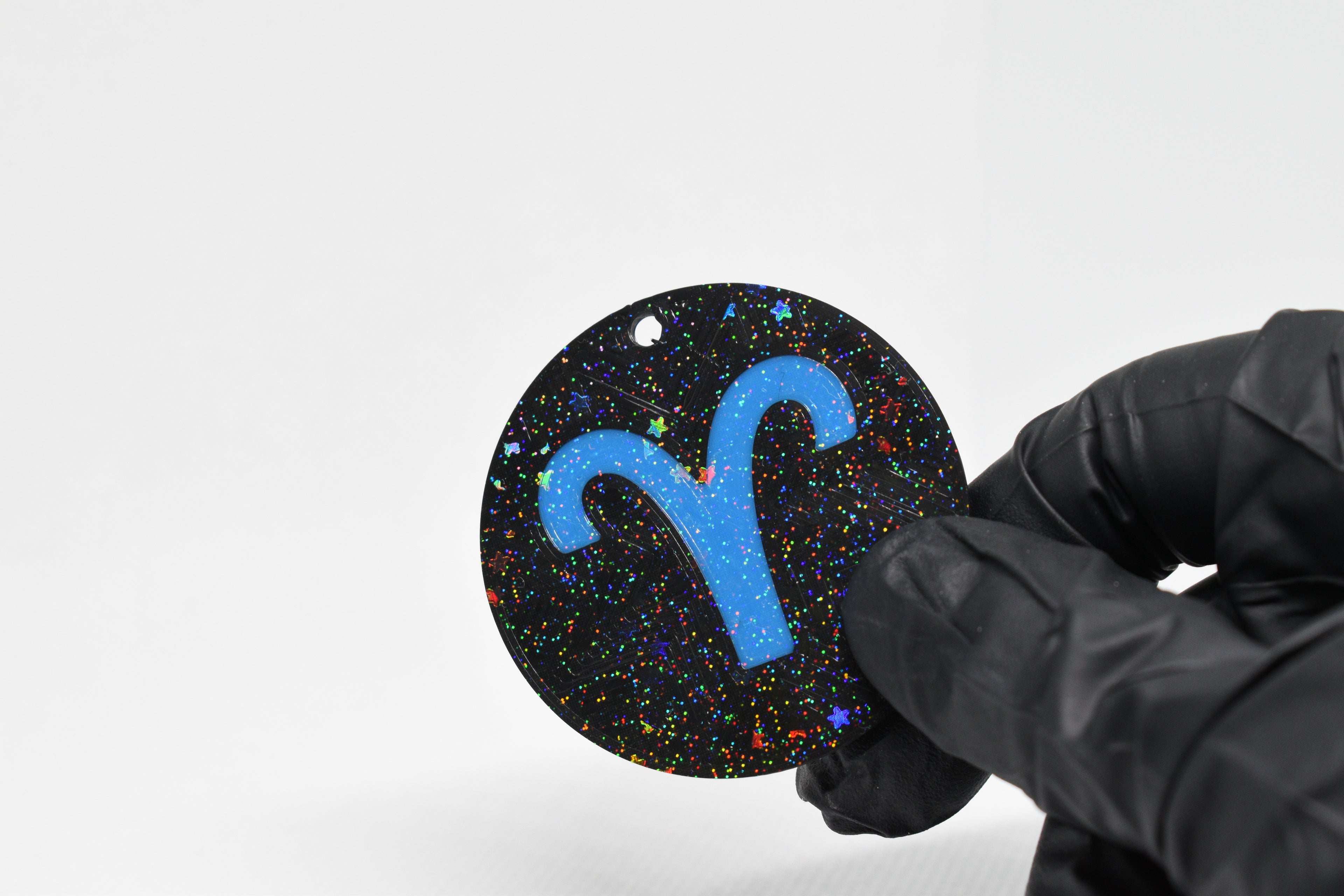 Aries zodiac keychain with blue symbol and glitter‑black cosmic background held in a gloved hand.