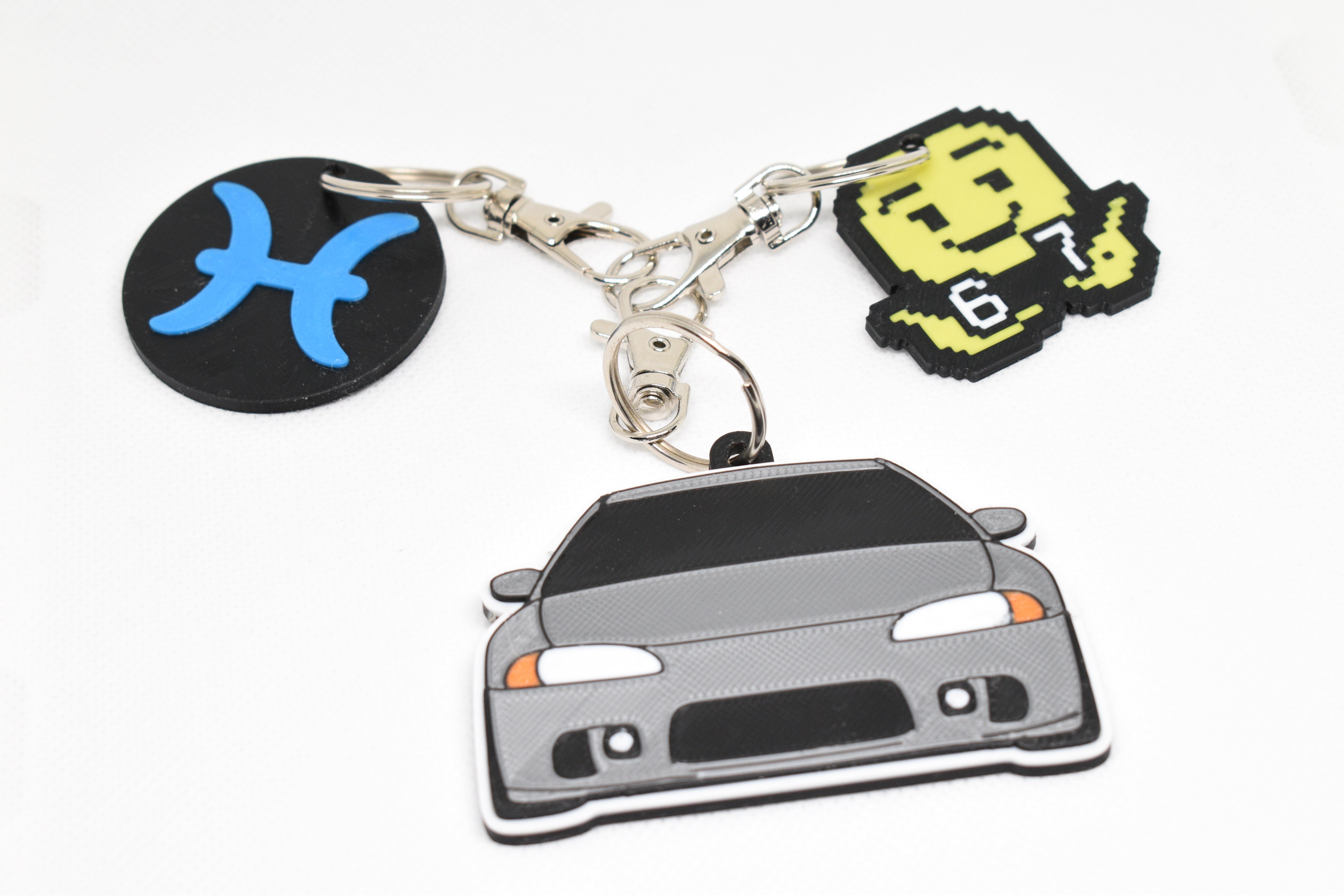Pisces zodiac keychain, pixel 67 emoji keychain, and gray Eclipse keychain attached together on metal rings.
