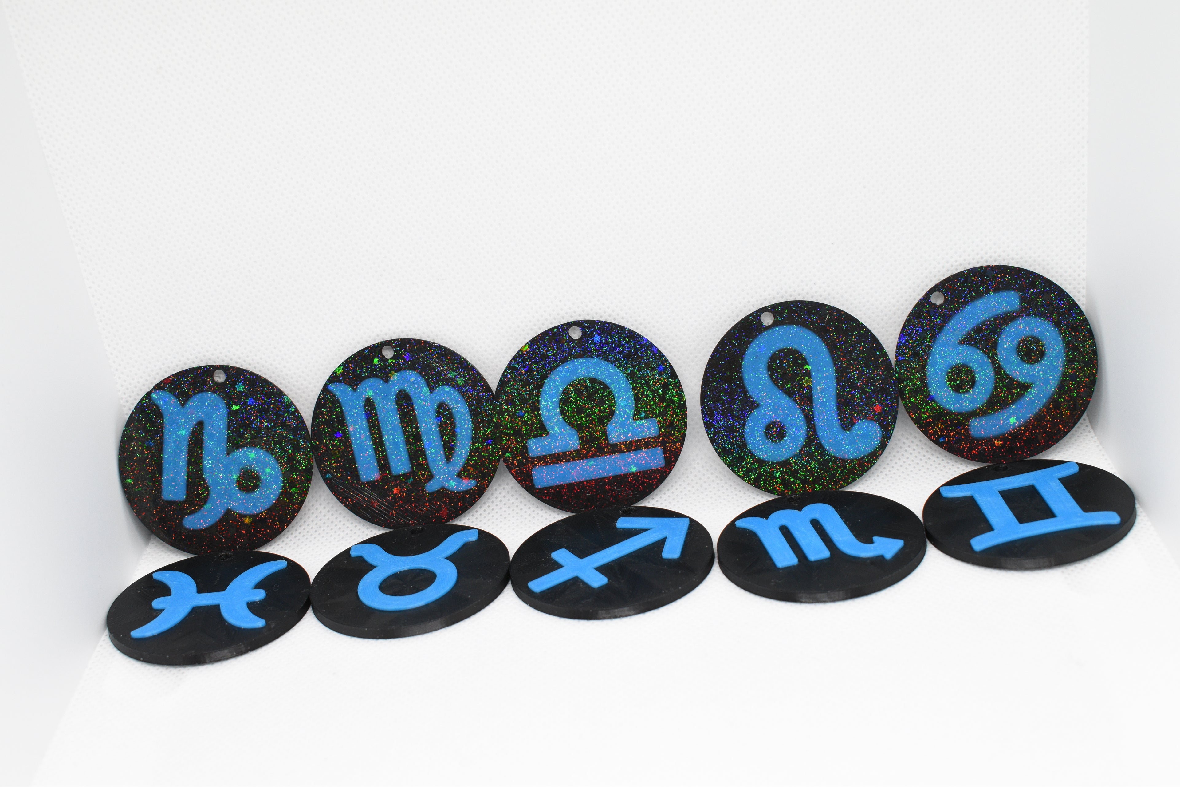 Set of glitter‑black zodiac keychains with bright blue astrology symbols arranged in two rows on a white background.