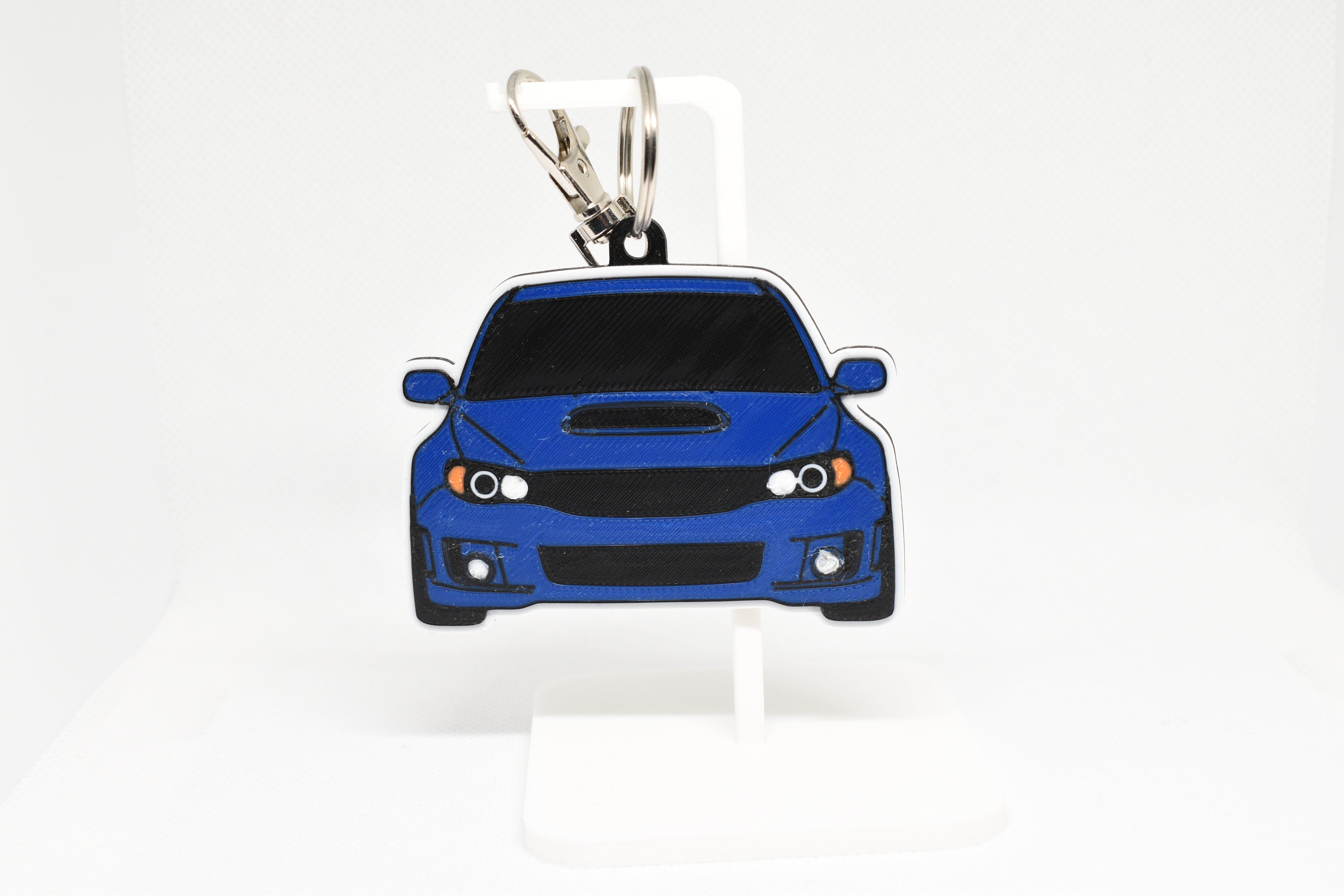 Blue WRX STI GR keychain displayed on a white stand with metal ring and clip.