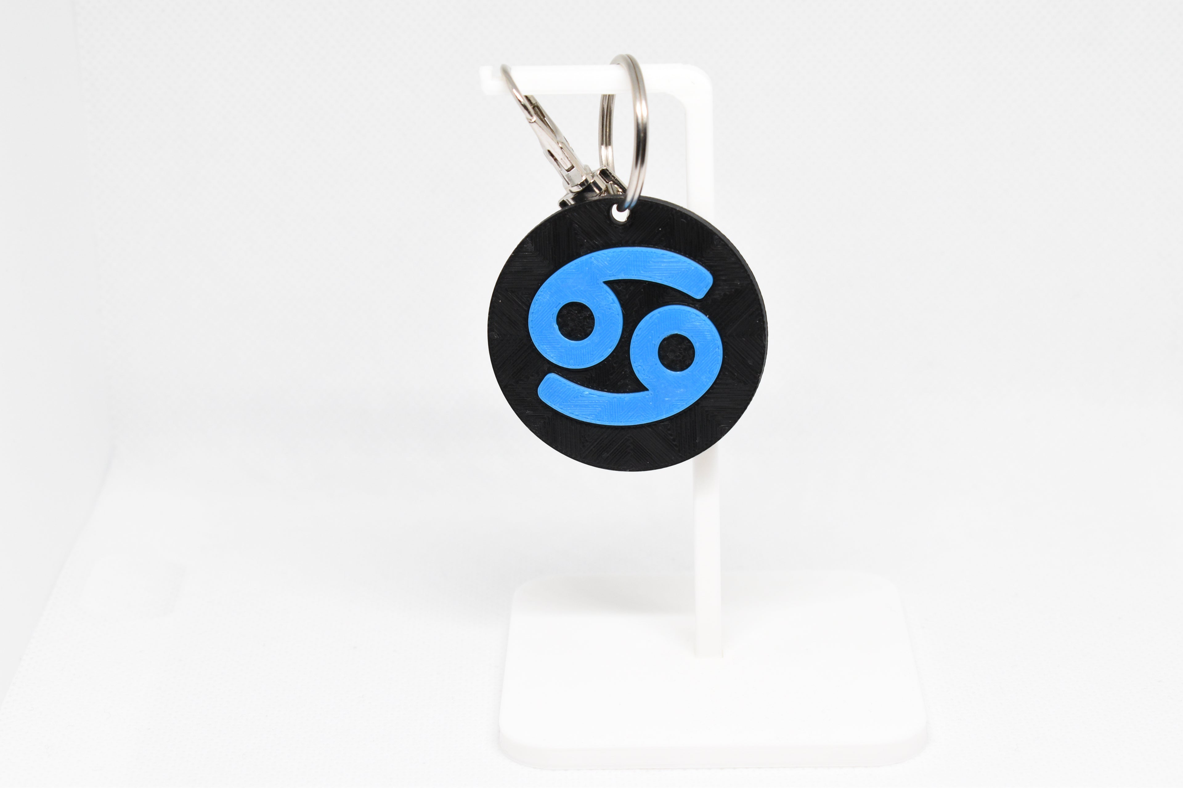 Cancer zodiac keychain with blue symbol displayed on a white stand with metal ring and clip.