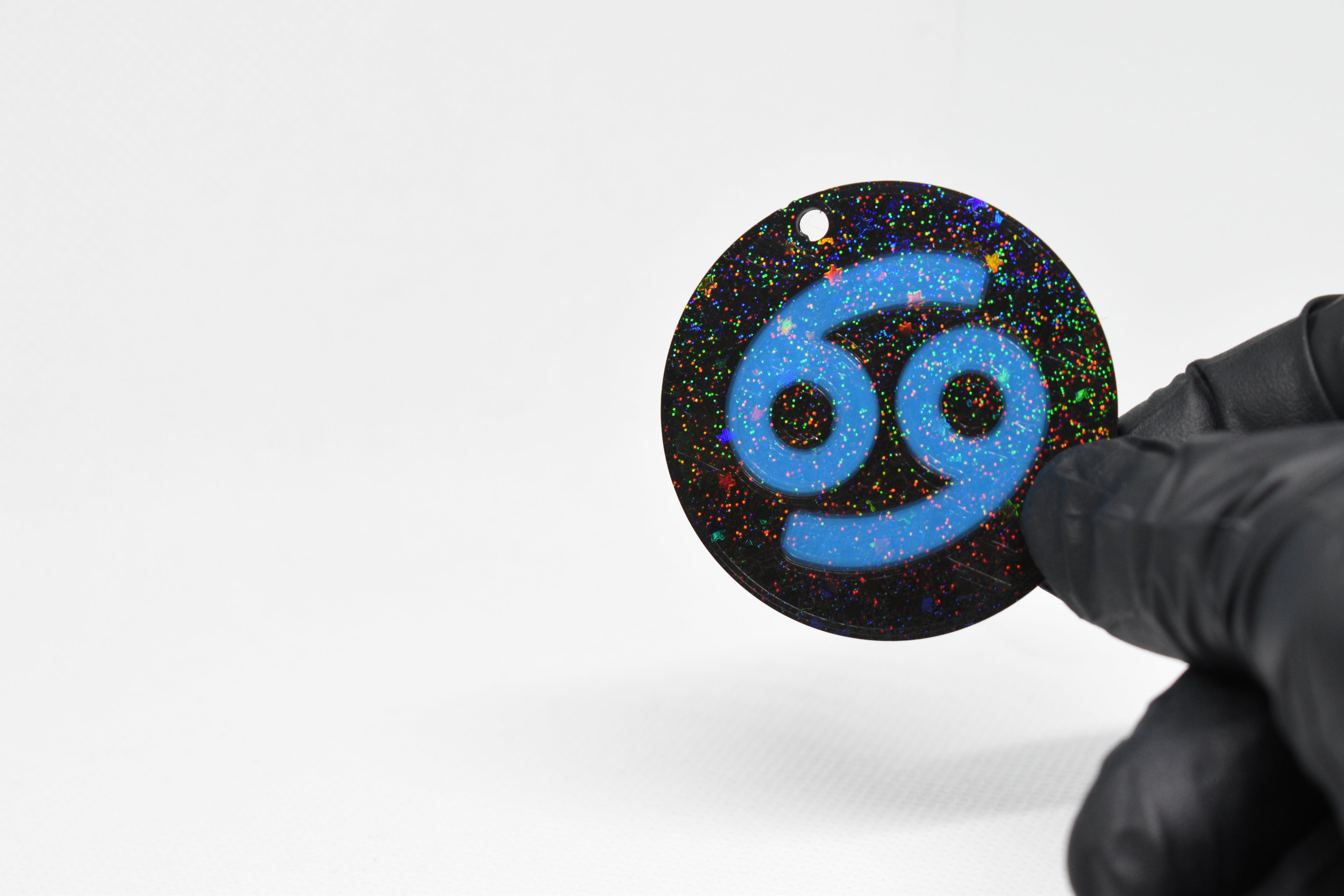 Cancer zodiac keychain with blue symbol and glitter‑black cosmic background held in a gloved hand.