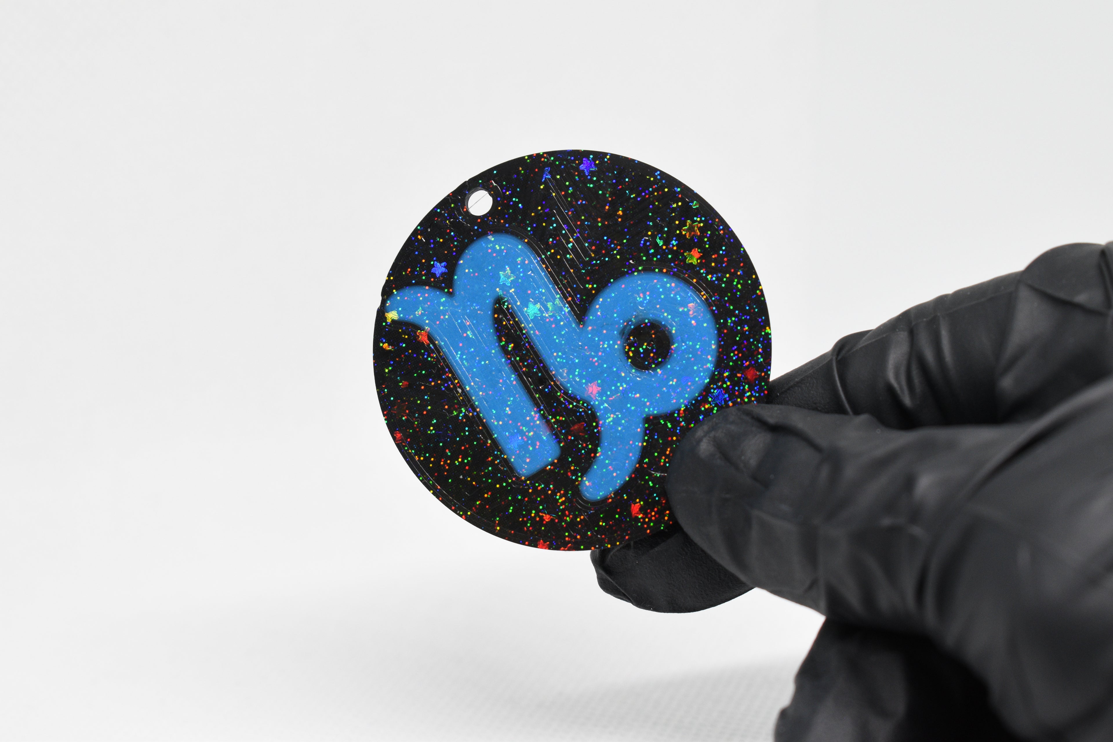 Capricorn zodiac keychain with blue symbol and glitter‑black cosmic background held in a gloved hand.