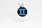 Gemini zodiac keychain with blue symbol displayed on a white stand with metal ring and clip.