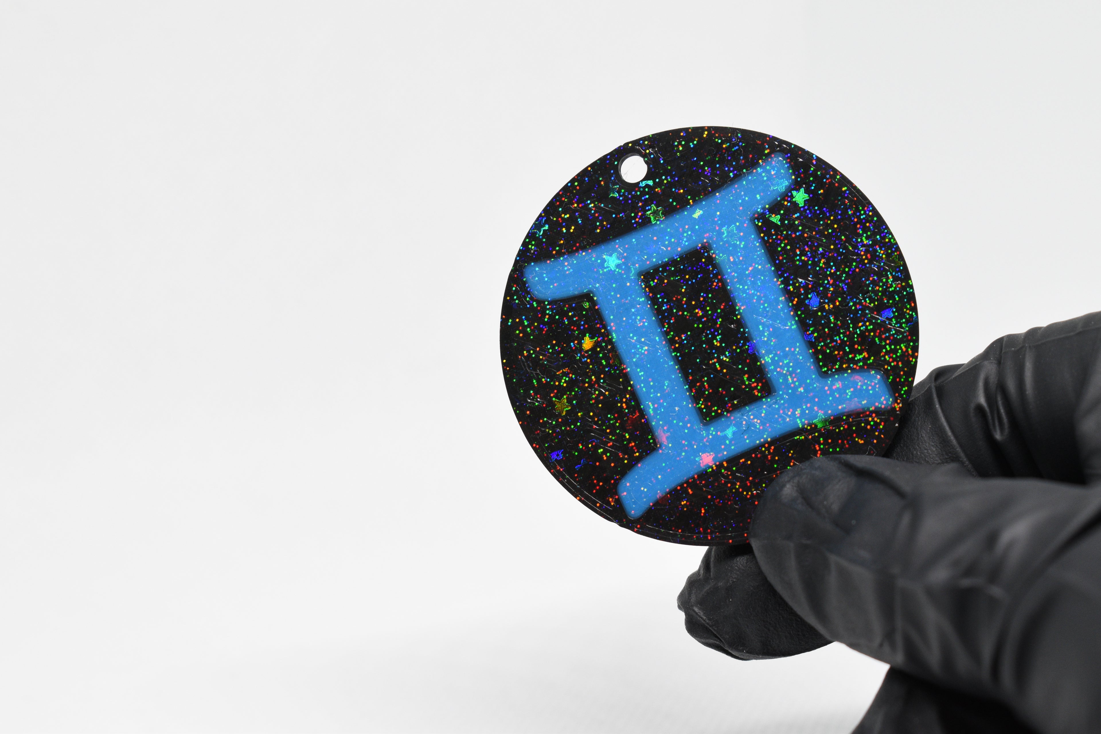 Gemini zodiac keychain with blue symbol and glitter‑black cosmic background held in a gloved hand.