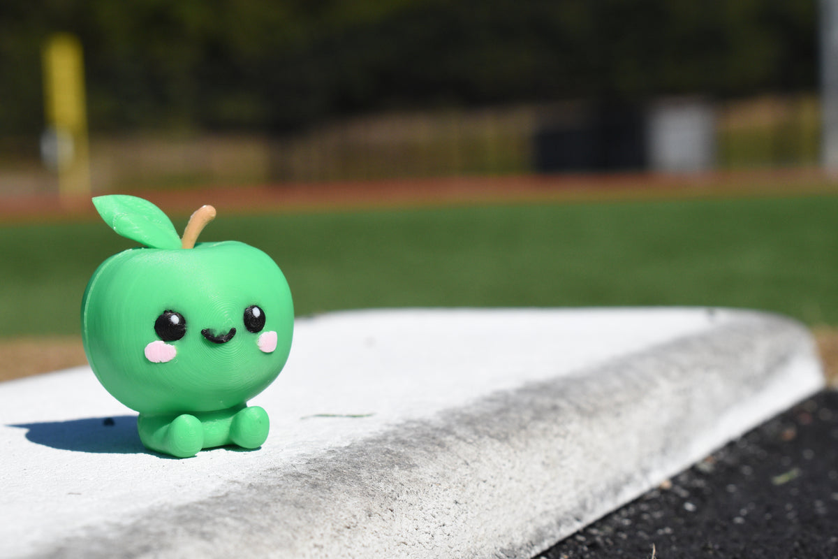 Cute 3D printed green kawaii green apple desk toy sitting outdoors on a white ledge with a sports field background by LayerPlay