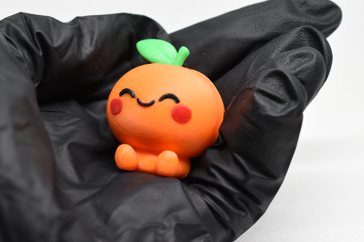 Hand in black glove holding cute 3D printed orange kawaii orange desk toy figurine close up by LayerPlay