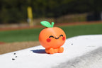 Cute 3D printed orange kawaii orange desk toy sitting outdoors on a white ledge with a sports field background by LayerPlay