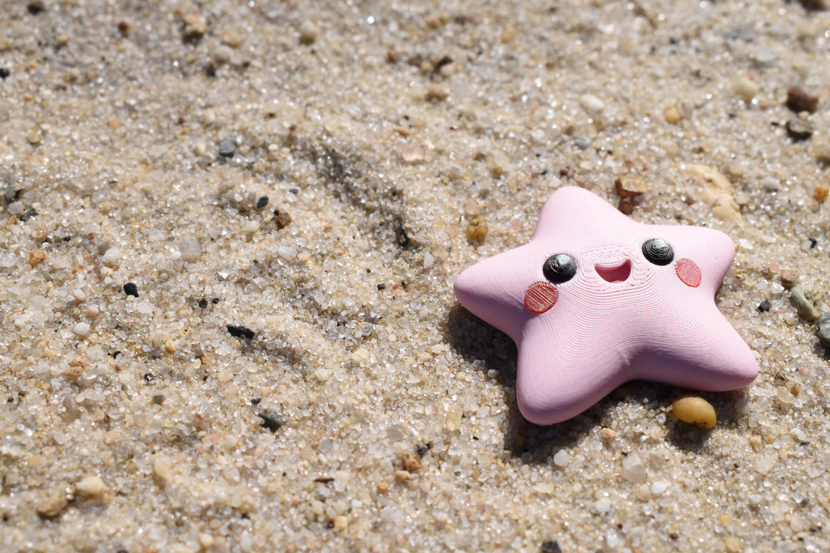 Pink 3D printed kawaii starfish desk toy figurine resting outdoors on beach sand by LayerPlay