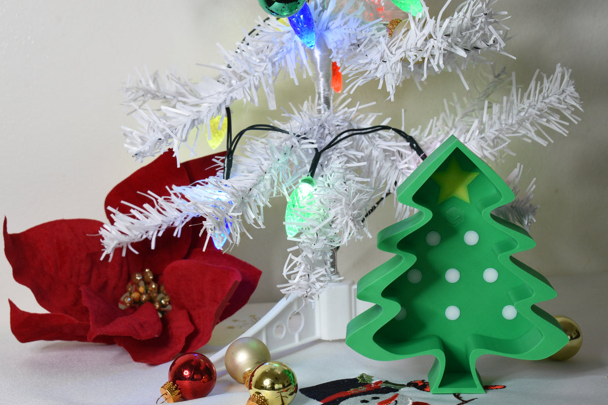 Layertreat FestiveTree green 3D printed Christmas tree candy dish displayed beside a red poinsettia flower, gold ornaments, and a lit white holiday tree.