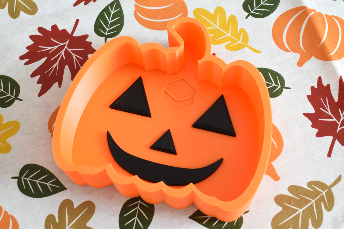 3D printed orange jack-o'-lantern candy dish displayed on a fall patterned tablecloth with leaf and pumpkin print by LayerPlay Layertreat