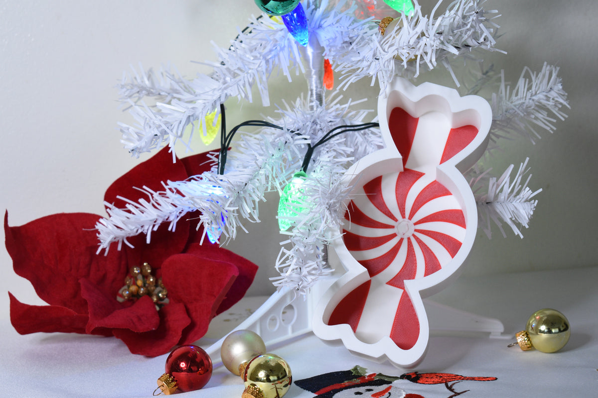 Layertreat MintyBowl red and white peppermint swirl candy dish displayed upright beside a white Christmas tree, red poinsettia flower, and shiny ornaments.