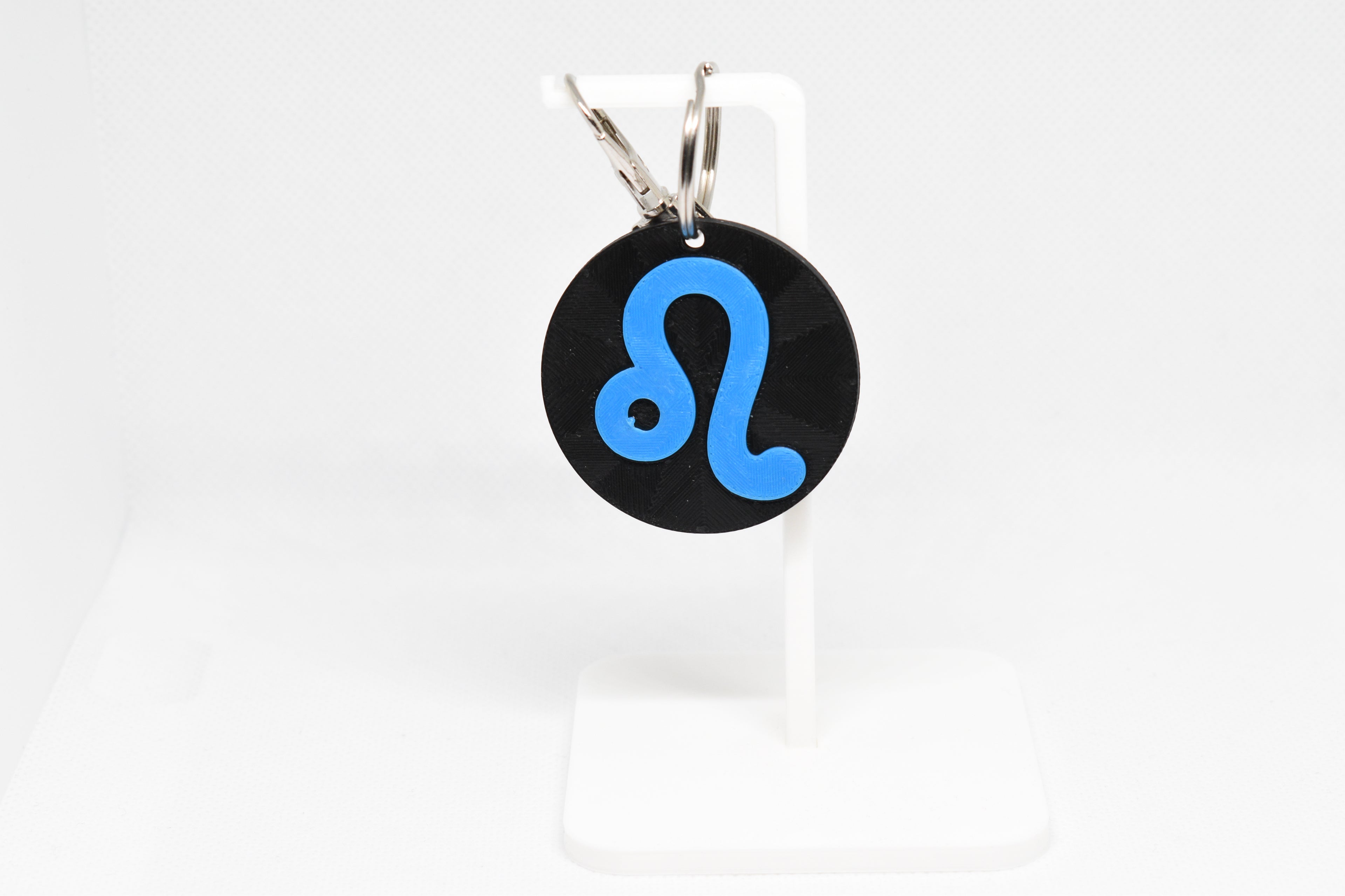 Leo zodiac keychain with blue symbol displayed on a white stand with metal ring and clip.
