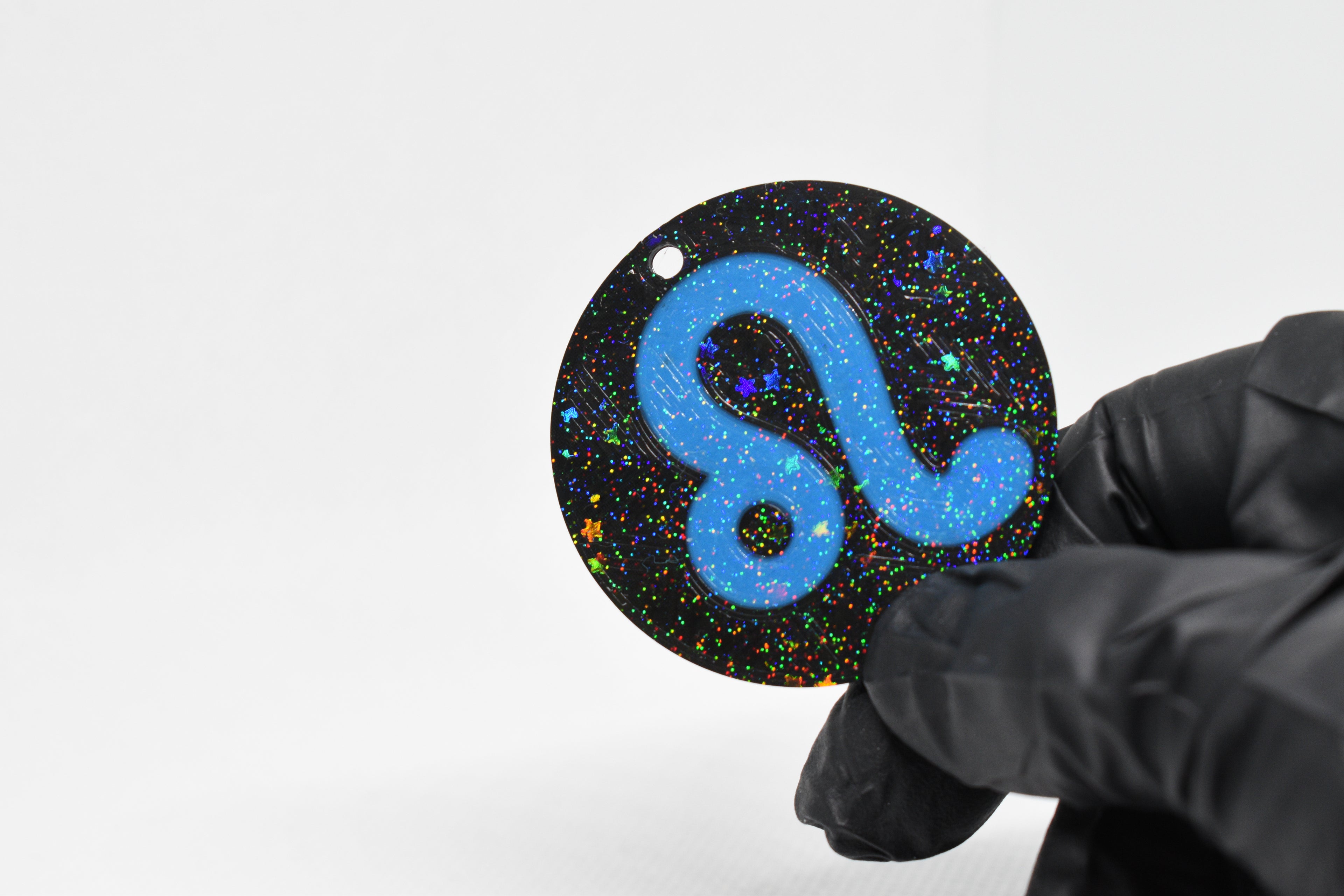 Leo zodiac keychain with blue symbol and glitter‑black cosmic background held in a gloved hand.