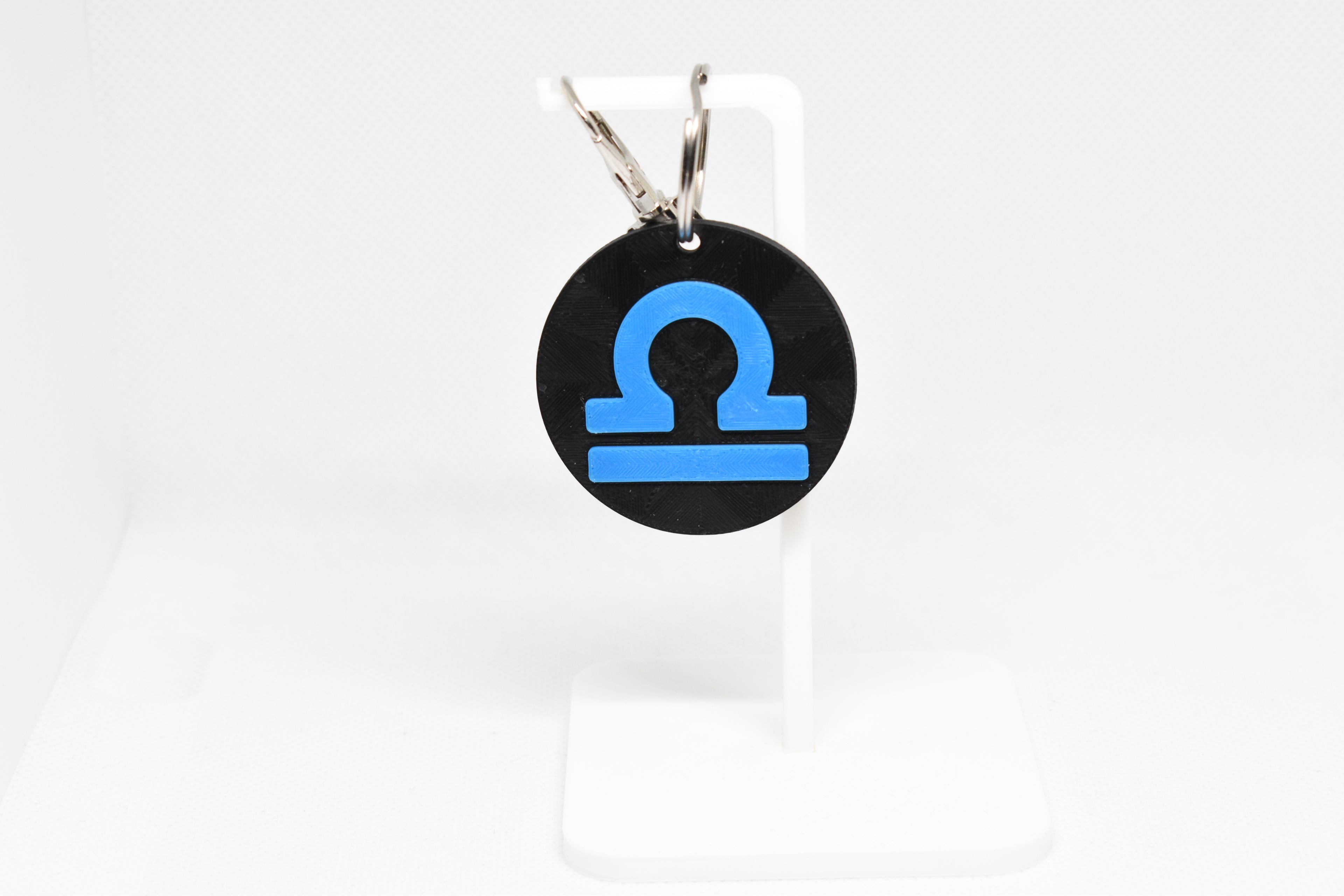 Libra zodiac keychain with blue symbol displayed on a white stand with metal ring and clip.