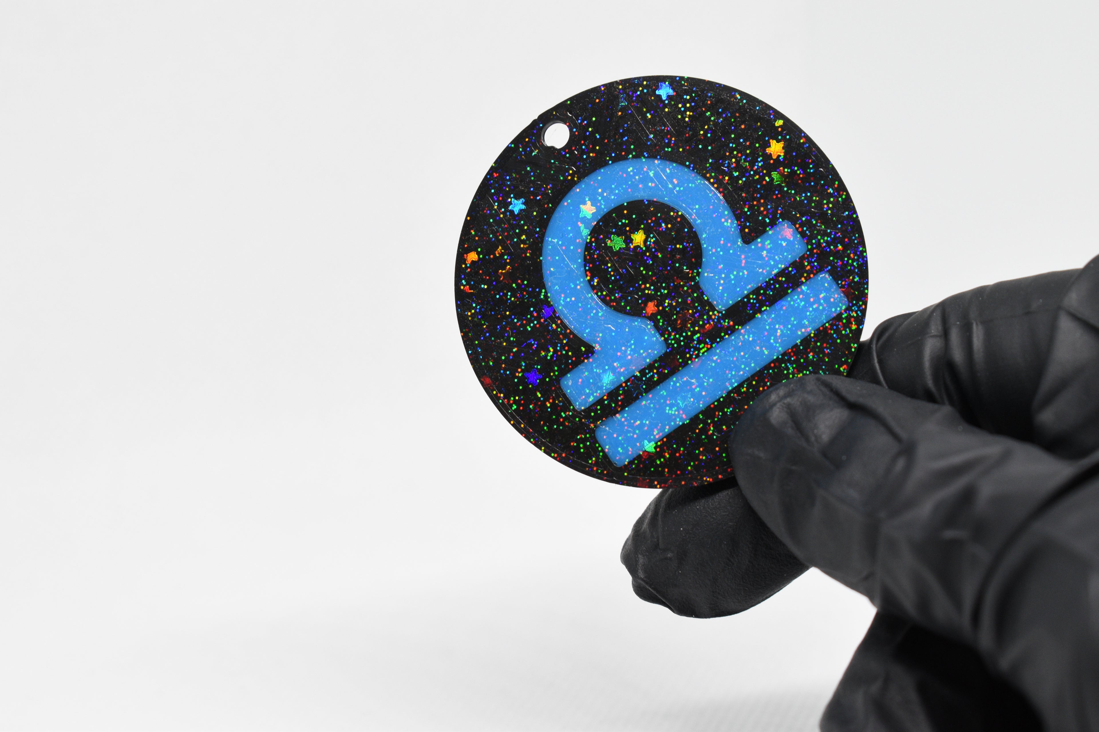 Libra zodiac keychain with blue symbol and glitter‑black cosmic background held in a gloved hand.