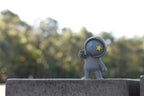 3D printed grey monomoodz drift desk figure standing outdoors on a stone ledge with blurred green trees in the background by LayerPlay