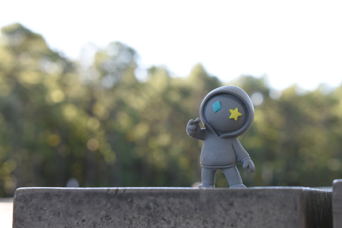 3D printed grey monomoodz drift desk figure standing outdoors on a stone ledge with blurred green trees in the background by LayerPlay