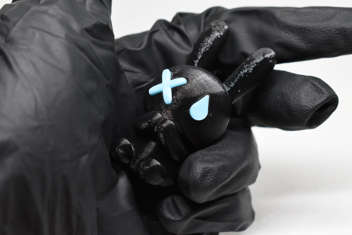 Hand in black glove holding 3D printed black monomoodz glow in the dark nixie bunny figure with white X and glow circle eyes close up by LayerPlay