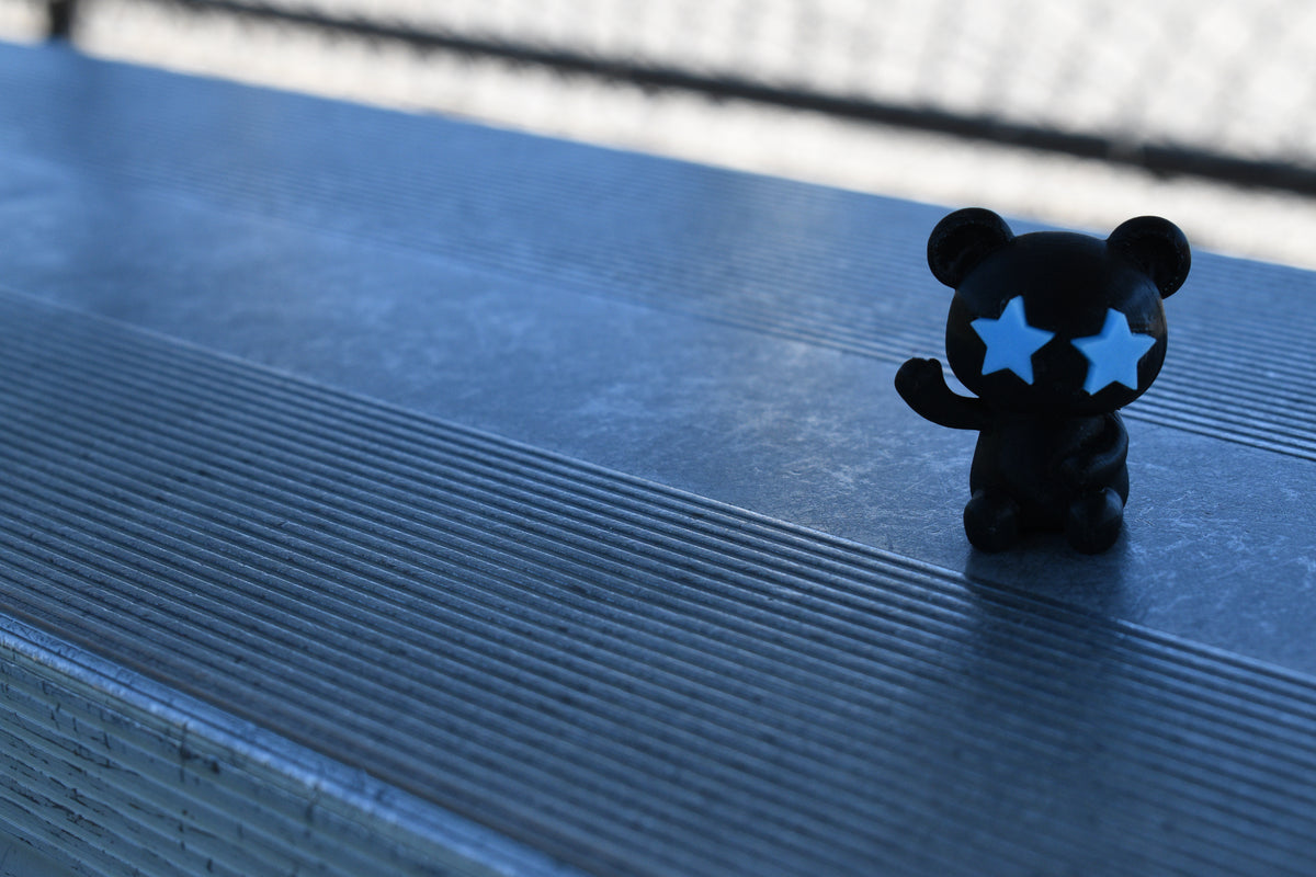 3D printed black monomoodz glow in the dark twinkle figure waving while sitting on metal stadium bleachers by LayerPlay