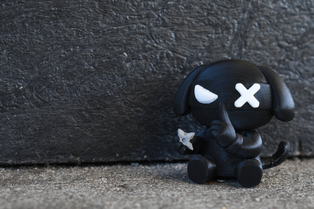 3D printed black monomoodz ninja glyphie dog desk figure sitting outdoors against a dark textured wall with shuriken in hand by LayerPlay