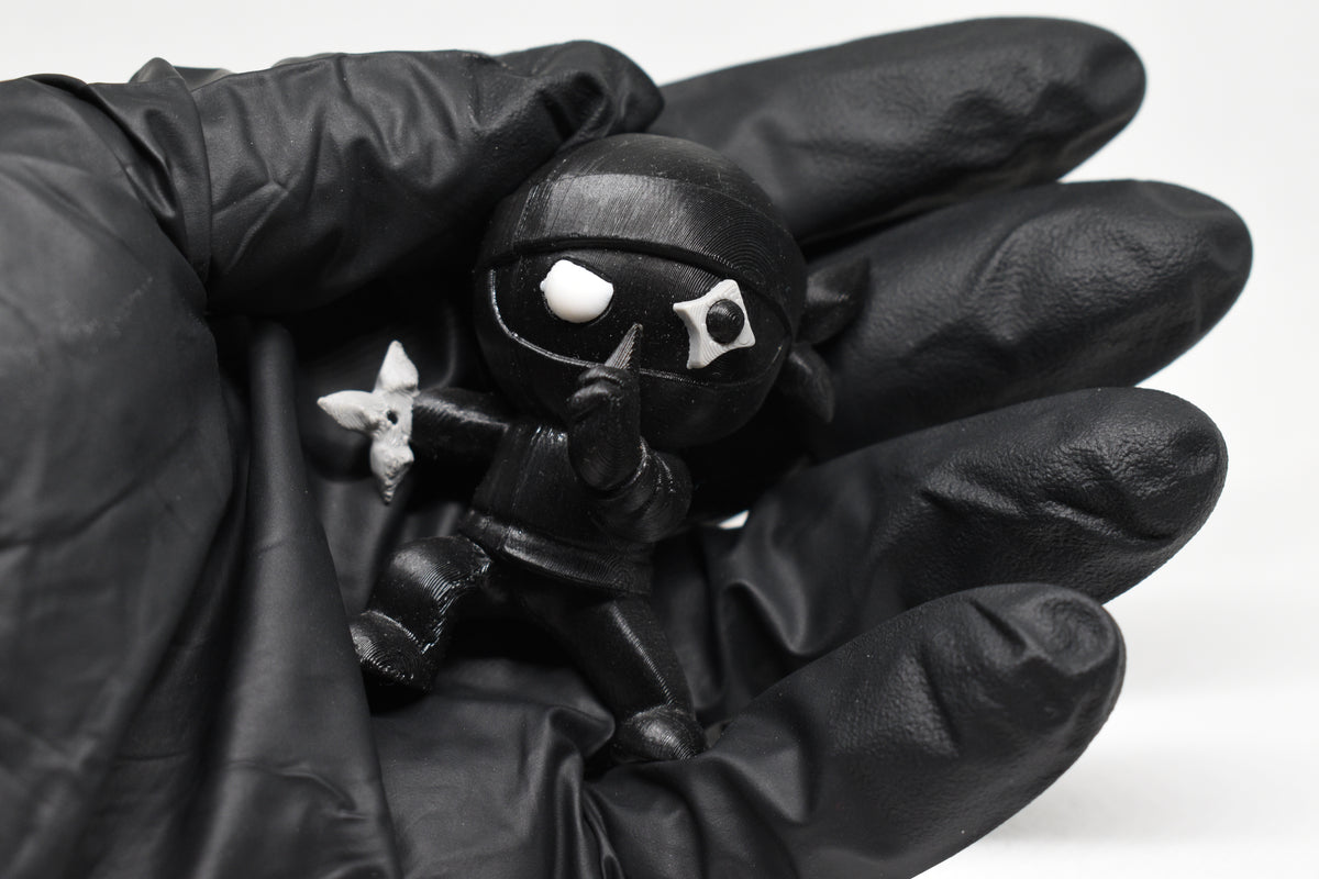 Hand in black glove holding 3D printed black monomoodz shurikid ninja desk figure close up by LayerPlay
