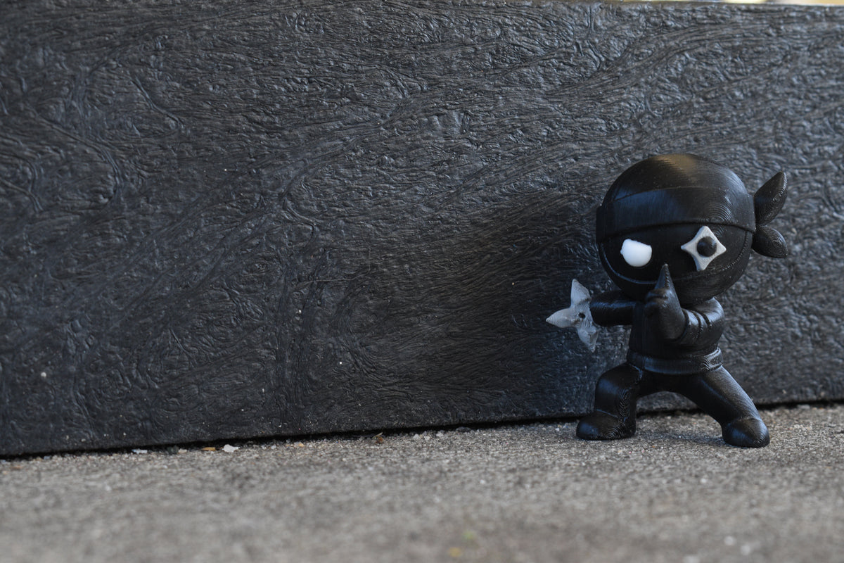 3D printed black monomoodz shurikid ninja desk figure posing stealthily outdoors against a textured dark background by LayerPlay