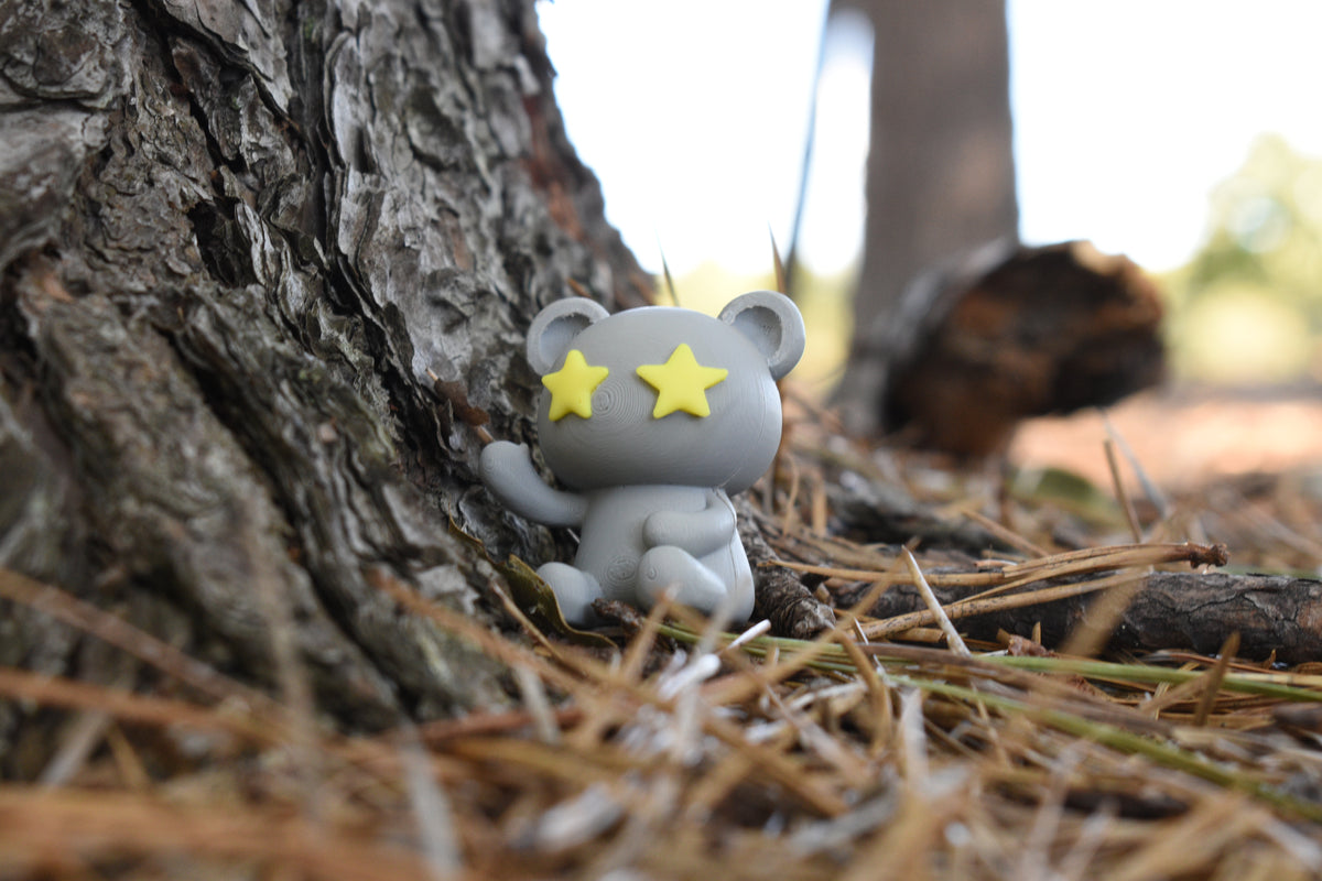 3D printed grey monomoodz twinkle desk figure sitting outdoors against a tree trunk on pine needles by LayerPlay