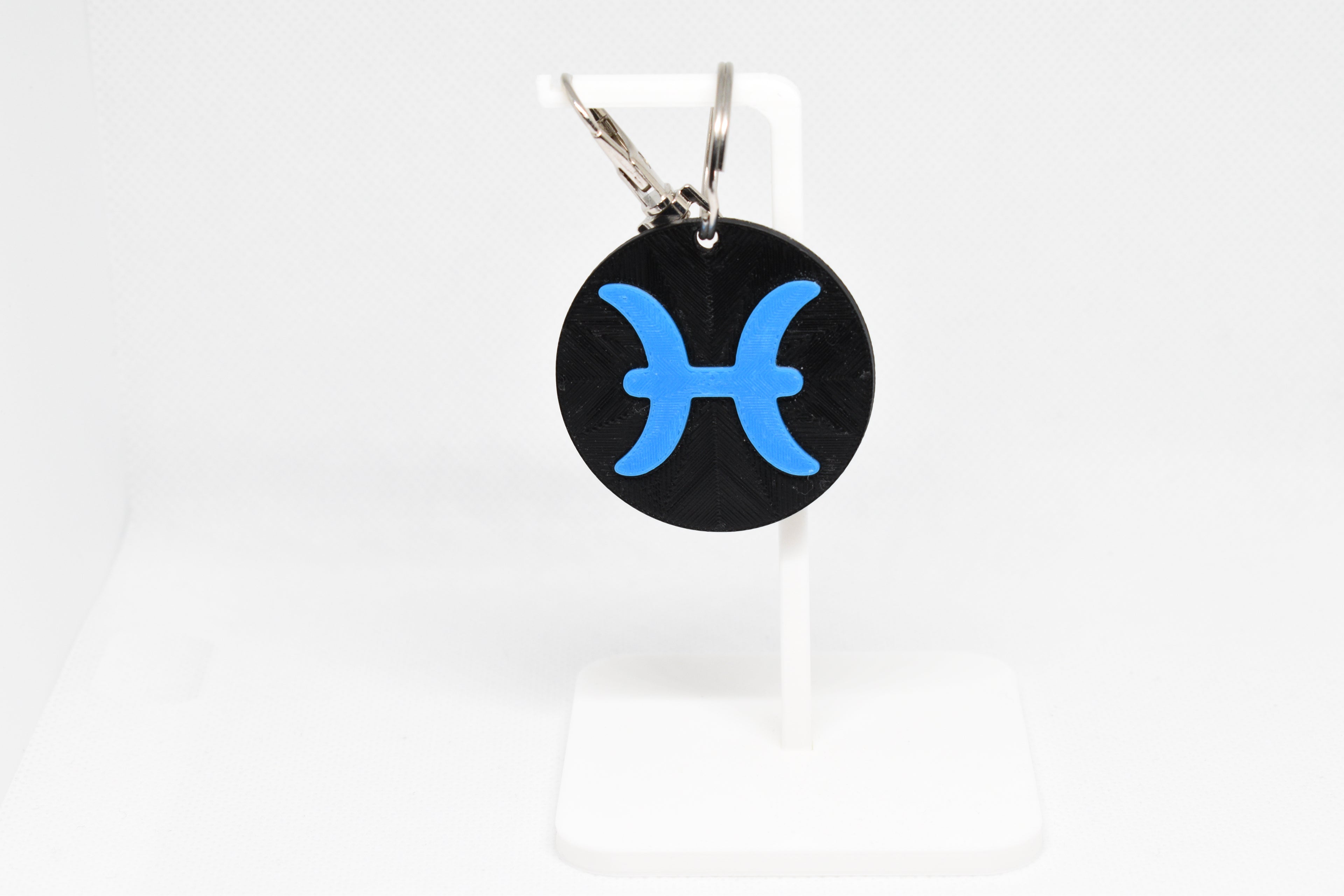Pisces zodiac keychain with blue symbol displayed on a white stand with metal ring and clip.