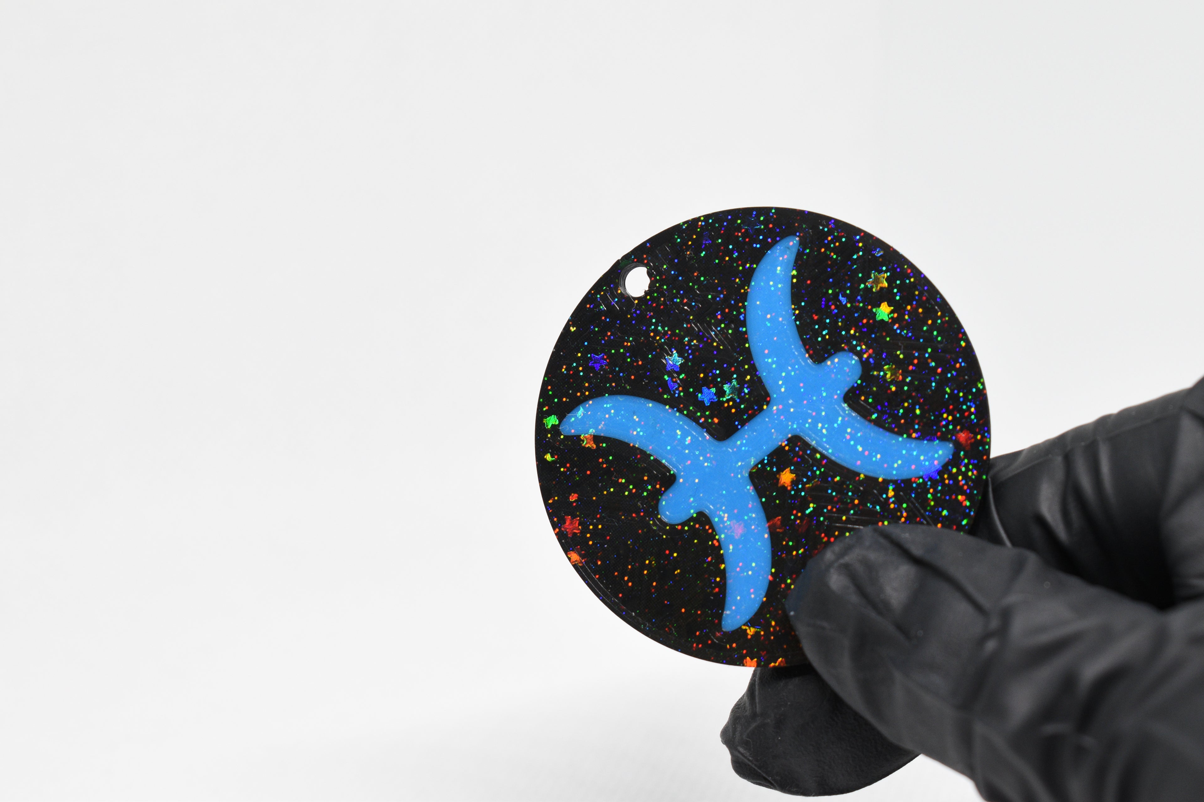 Pisces zodiac keychain with blue symbol and glitter‑black cosmic background held in a gloved hand.