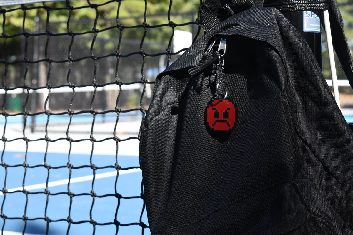 Red pixelated angry emoji keychain clipped to a black backpack zipper outdoors near a tennis court by LayerPlay