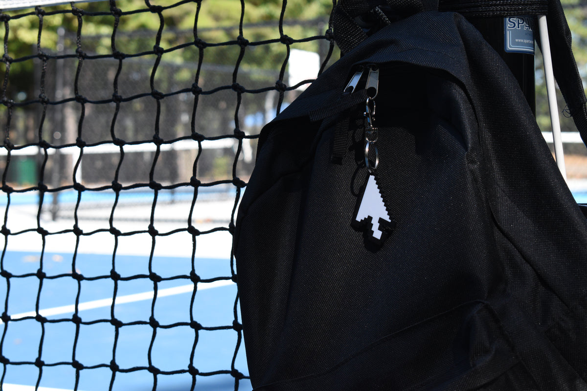 Pixelated arrow cursor keychain clipped to a black backpack zipper outdoors near a tennis court by LayerPlay