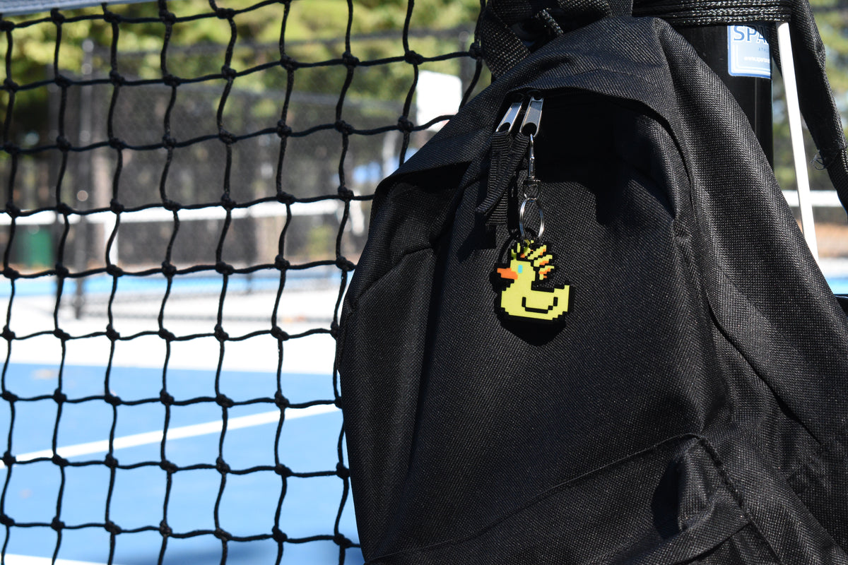 Ascended Duck pixel rubber duck keychain clipped to a black backpack zipper outdoors near a tennis court by LayerPlay