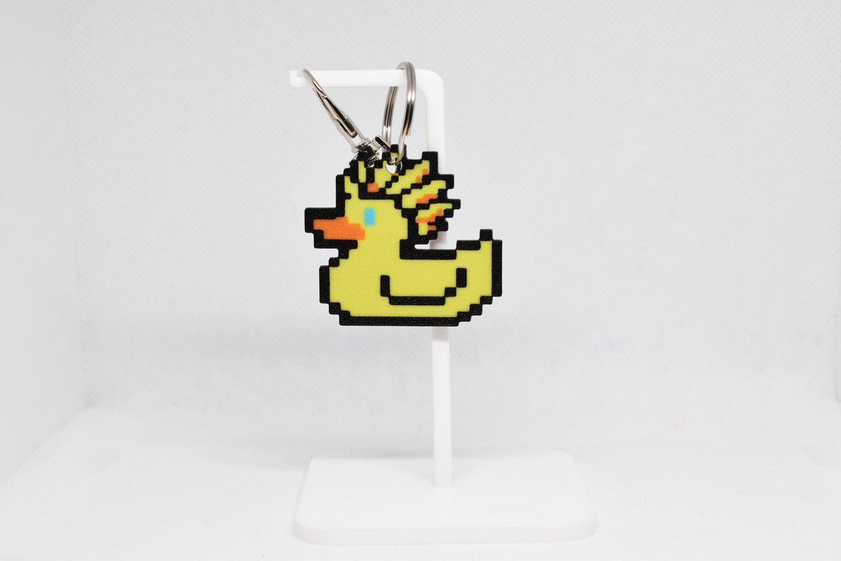 3D printed retro Ascended Duck pixel rubber duck keychain hanging on a white display stand by LayerPlay